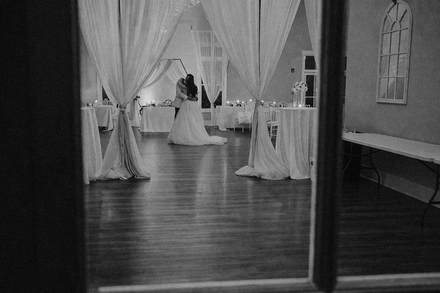 Black and white photo of a couple sharing a first dance in an elegant wedding reception hall with draped fabric columns.