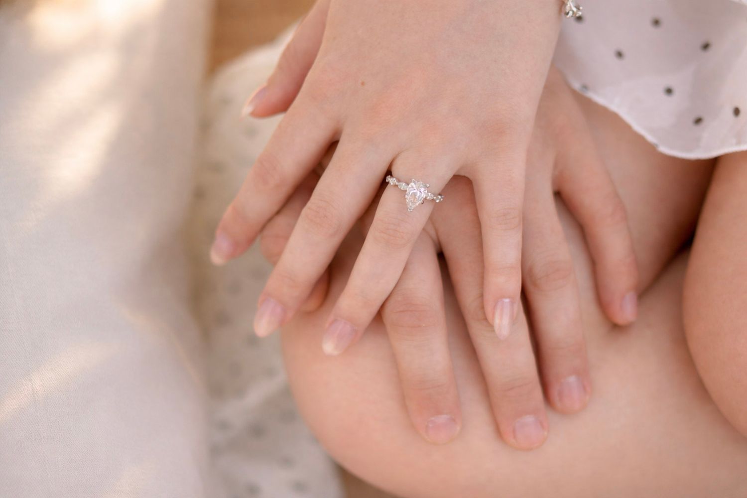 A delicate engagement ring sparkles on a woman's hand resting against light fabric.