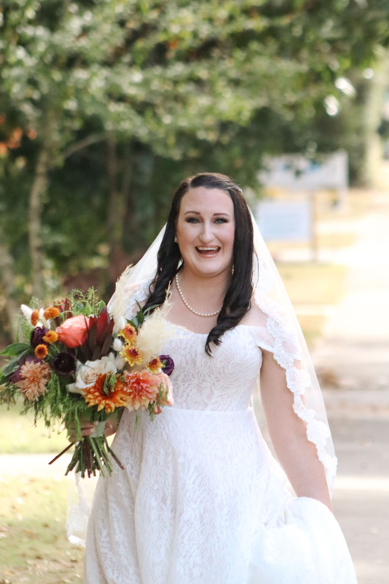 Bride in a white lace dress holds a vibrant fall-colored bouquet with orange and red flowers in an outdoor setting.