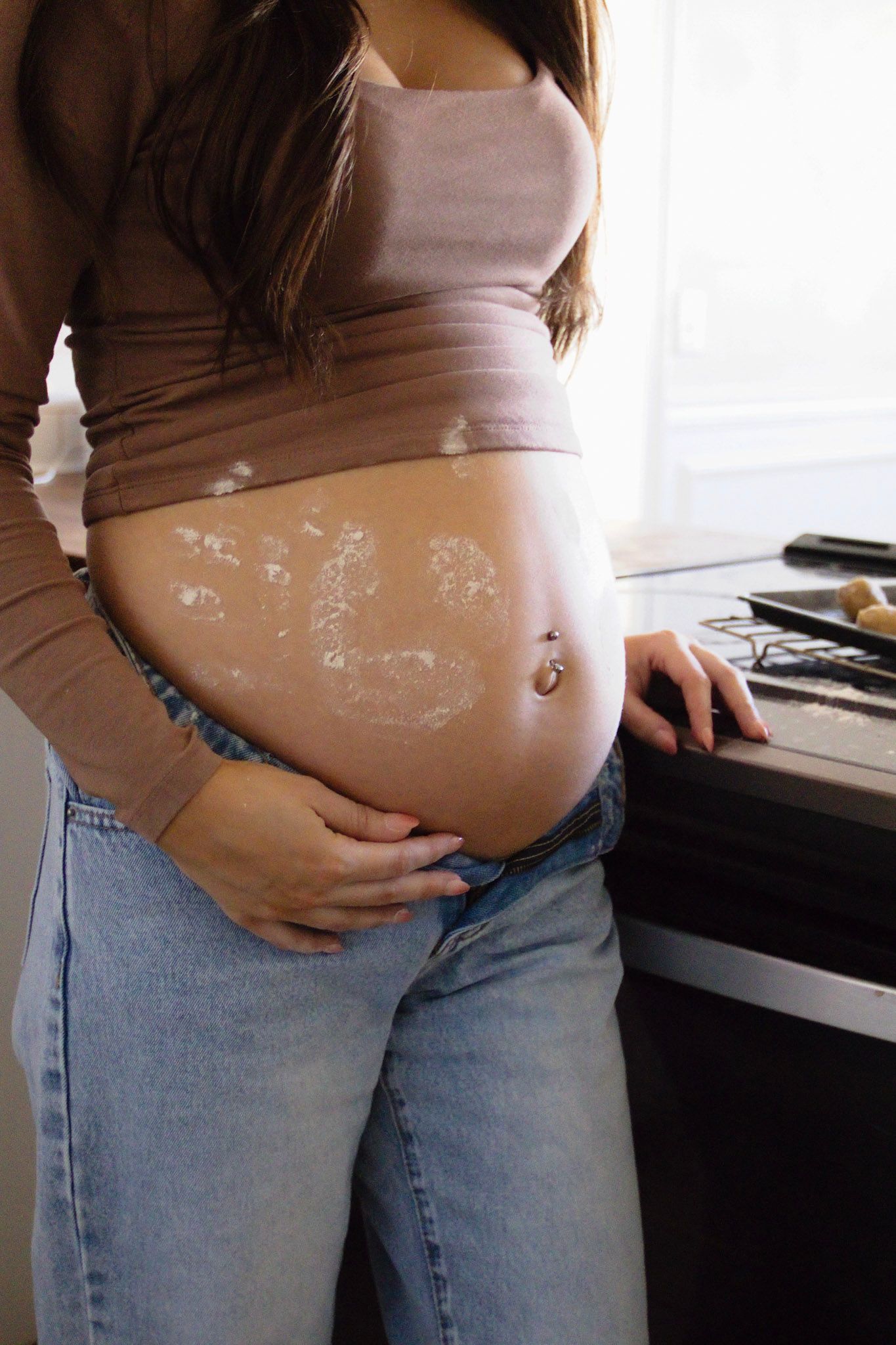 Pregnant woman in kitchen with flour on her baby bump, wearing jeans and a cropped top, baking during pregnancy.