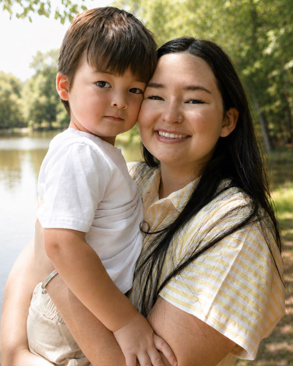 A smiling young woman holds a toddler boy outdoors near a lake surrounded by lush green trees.