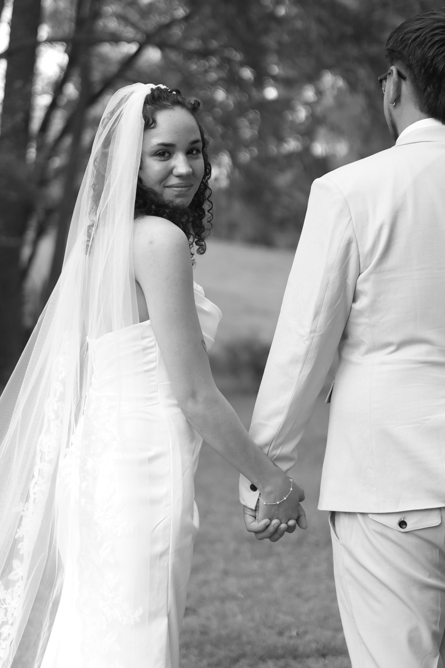 Black and white photo of a bride in a veil holding hands with a groom in a white suit outdoors.