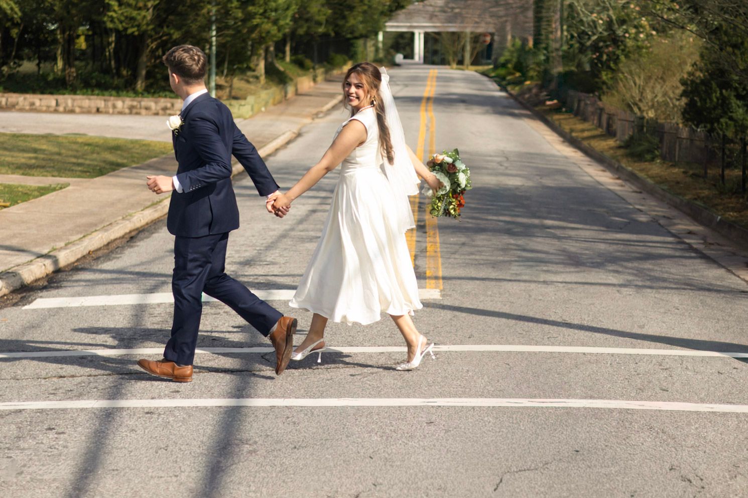 A couple in wedding attire playfully walks across a sunny suburban street holding hands.
