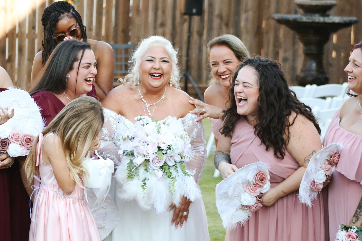 Wedding party laughing and celebrating in pink dresses against wooden fence backdrop.