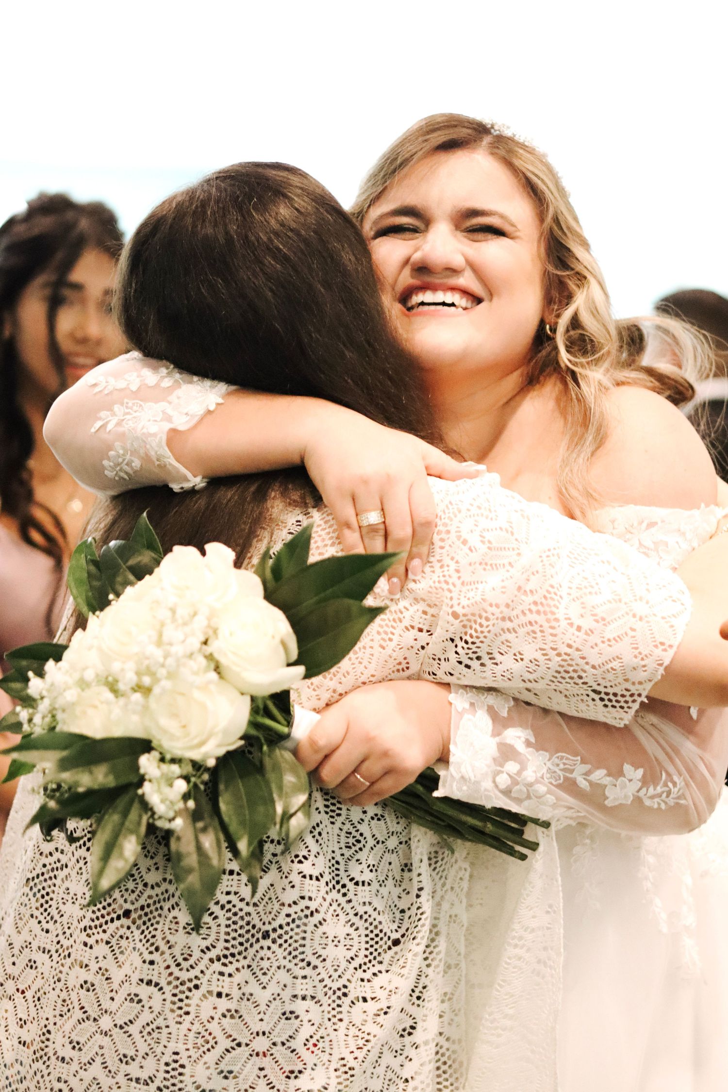 Wedding party members in white lace dresses embrace and smile while holding white floral bouquets.