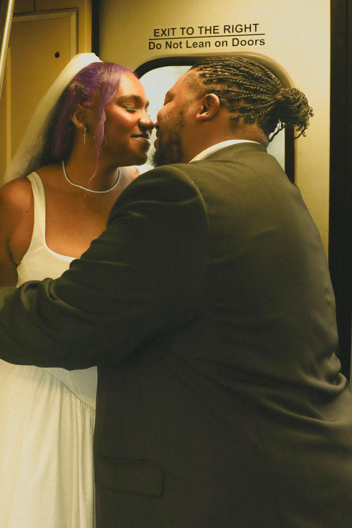 A bride and groom share a romantic kiss on a subway train, bride wearing white dress and veil with purple hair.