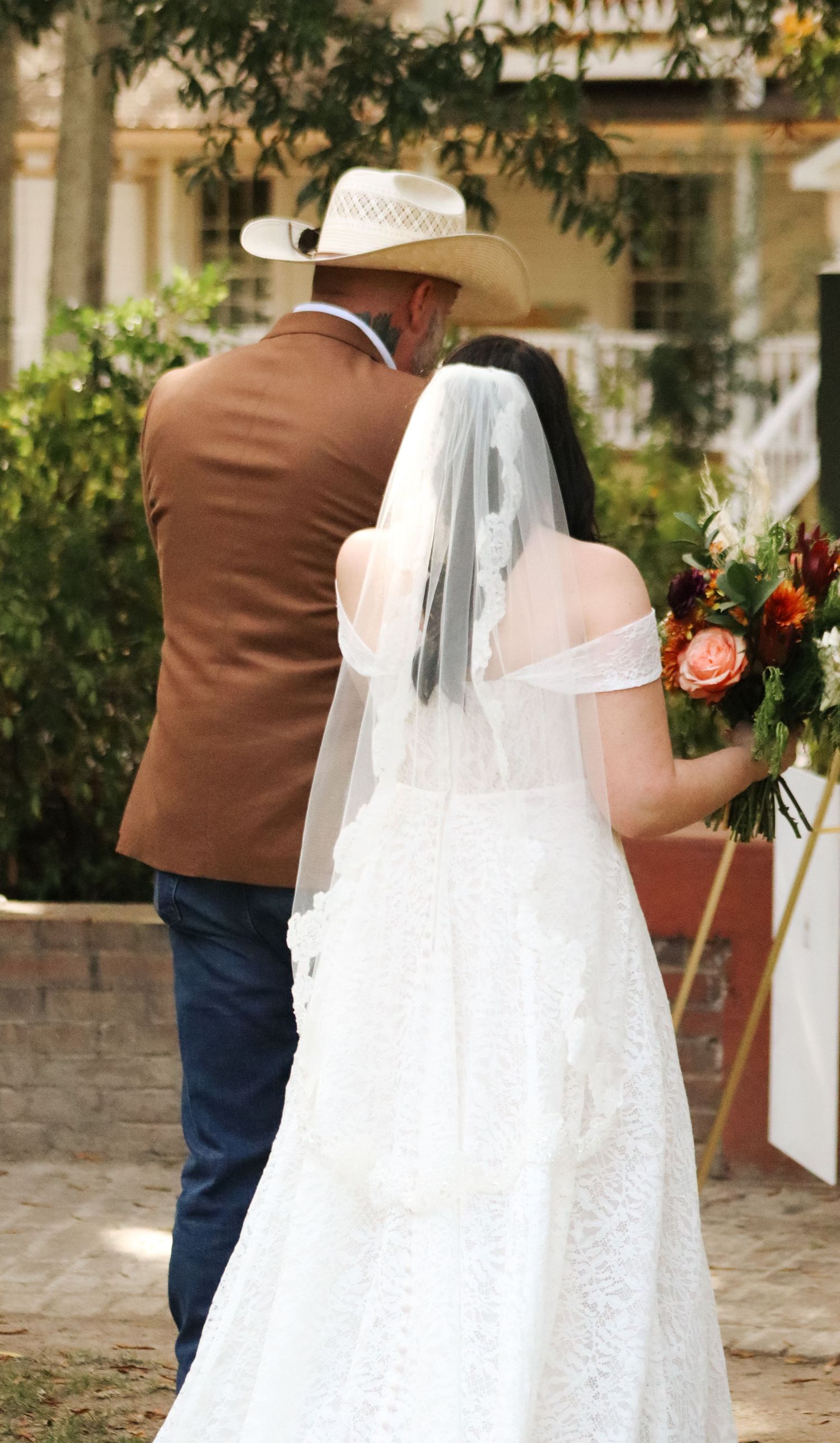 Wedding couple in brown suit and white lace dress with veil photographed from behind.