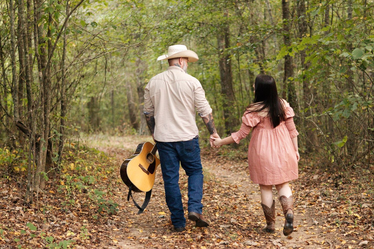 A couple walks hand in hand through autumn woods carrying a guitar.