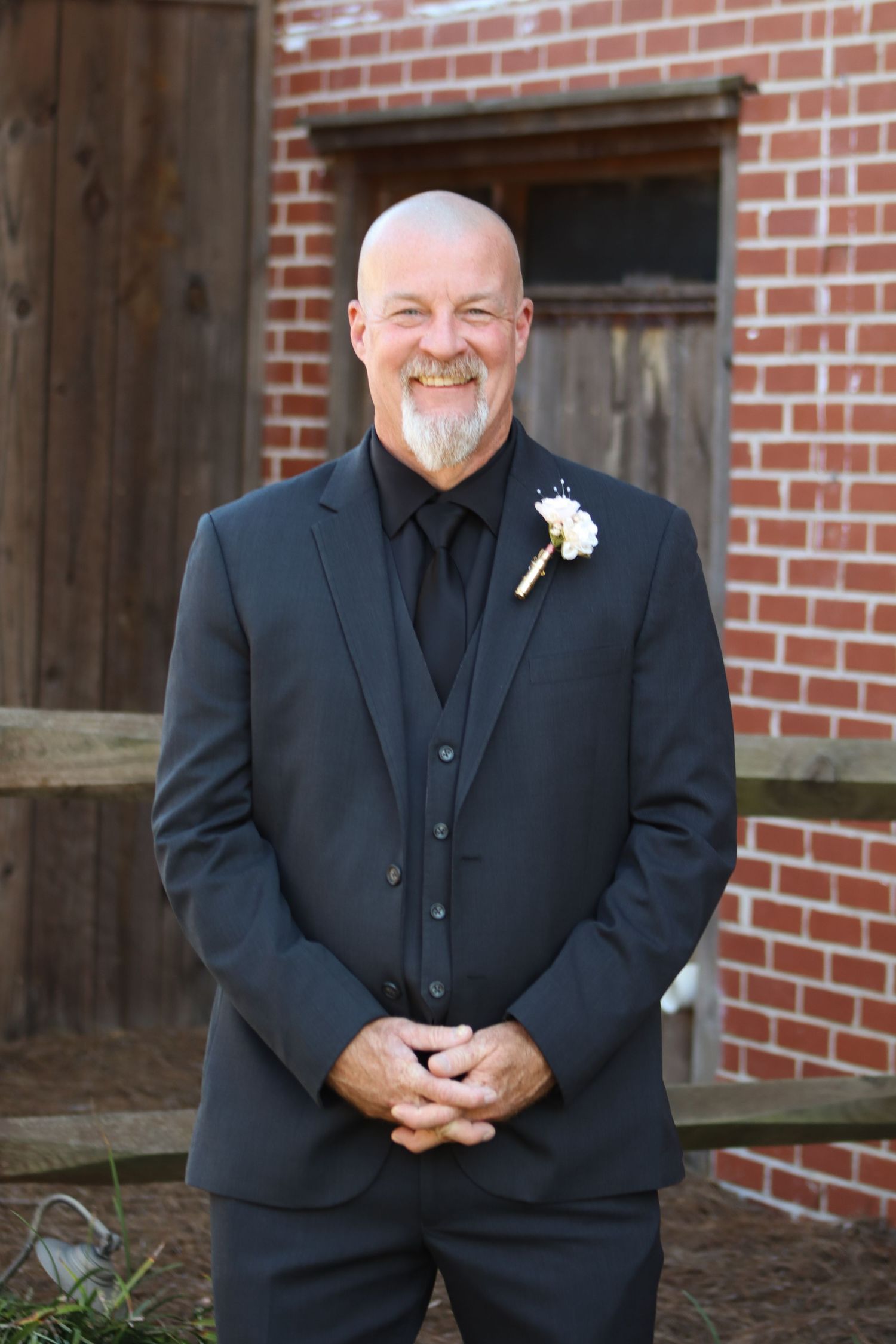 Groomsman in black suit and vest smiles warmly against brick wall background.