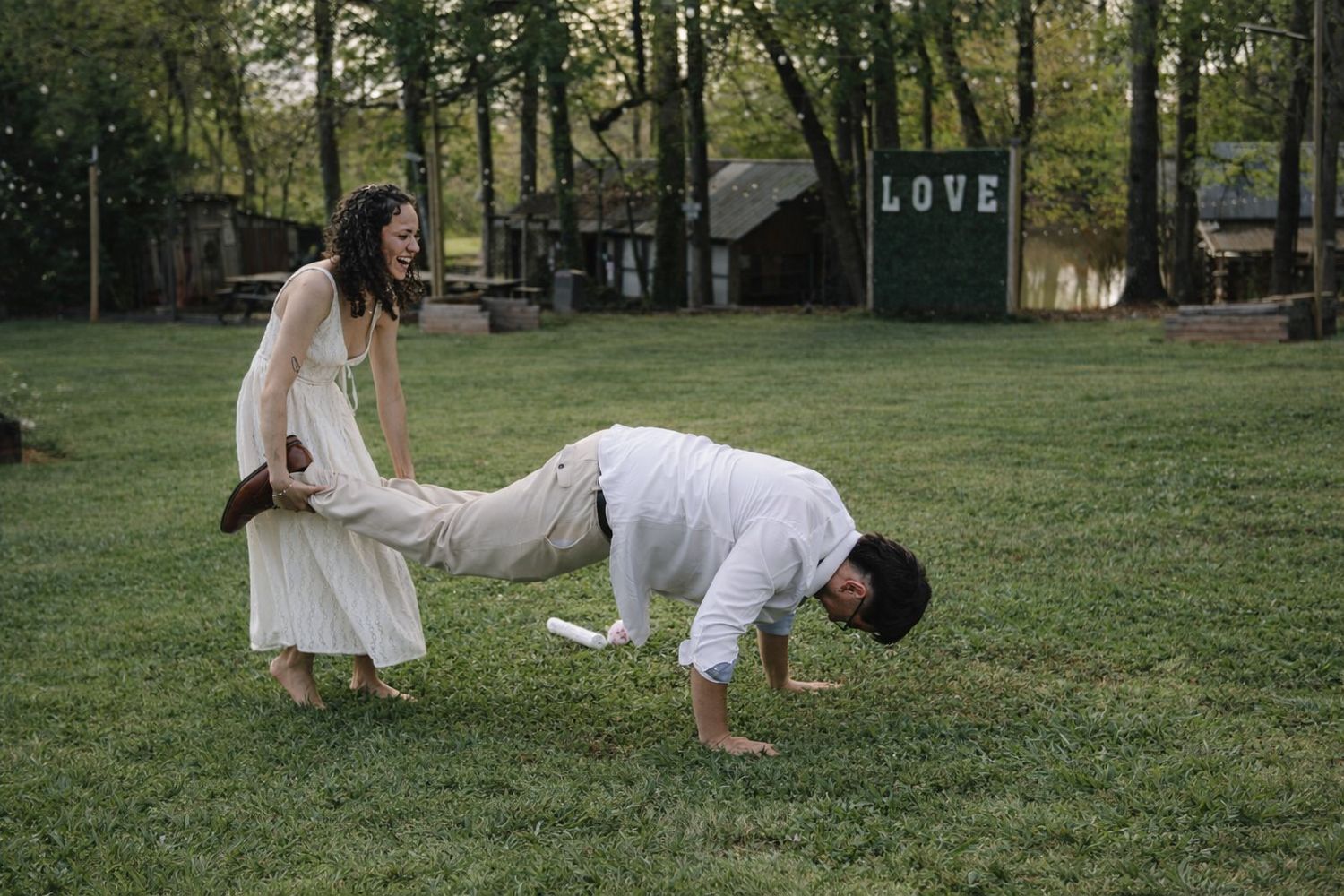 Laughing bride in white dress playfully pulls groom's leg on green lawn at outdoor wedding venue.