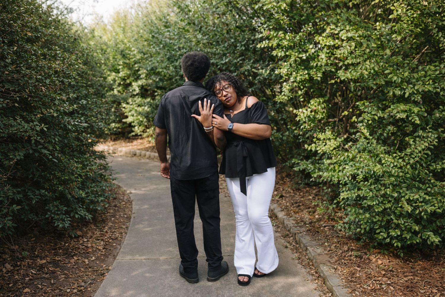 Two people in formal attire walk together down a tree-lined path during a special occasion.
