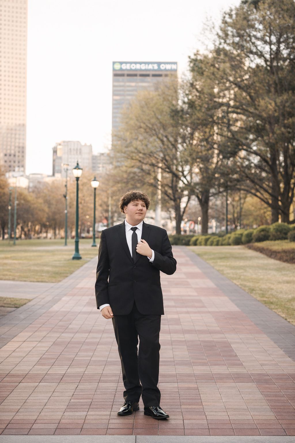 A figure in a black suit stands contemplatively on a paved walkway with trees and buildings in the background.