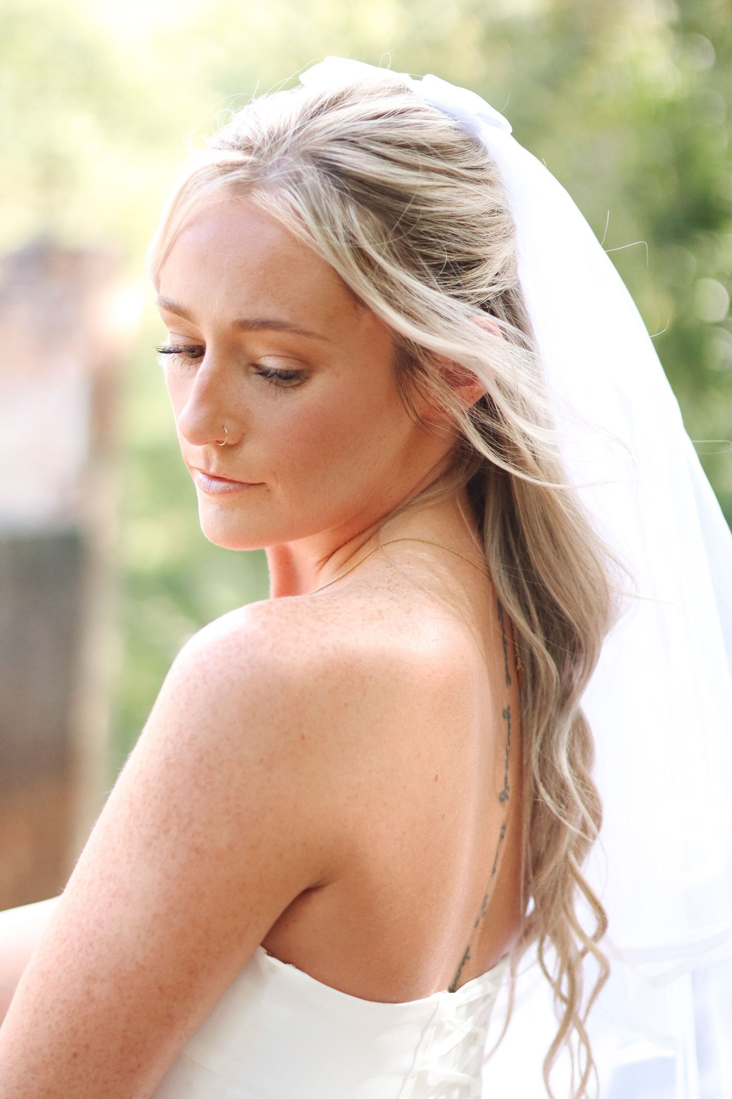 Side profile of a bride in a white dress with flowing blonde hair and veil against a soft natural background.