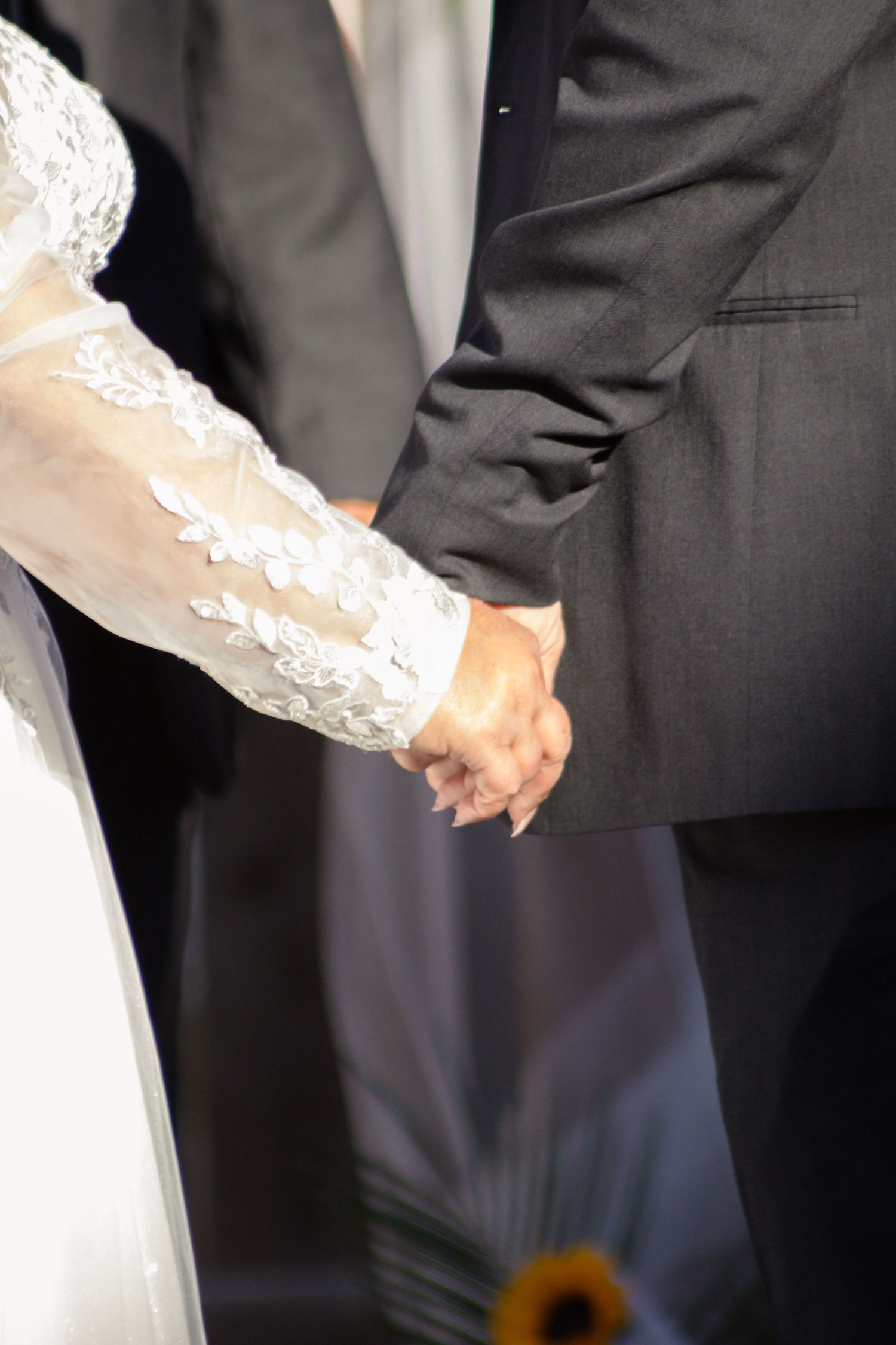 Close-up of bride and groom holding hands during wedding ceremony with white lace sleeve detail visible.