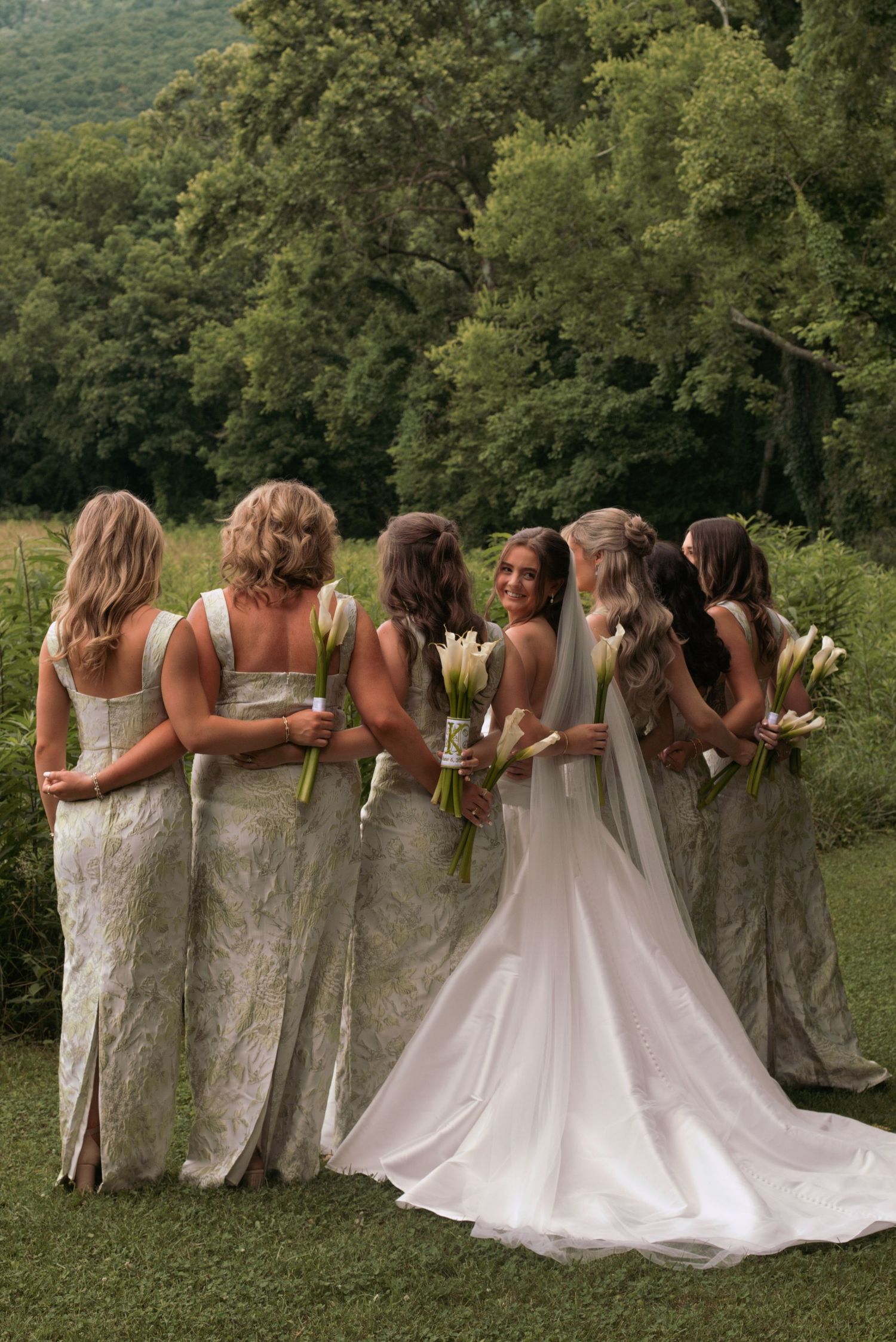 Bride in white gown smiles with bridesmaids in sage lace dresses holding white floral bouquets outdoors.