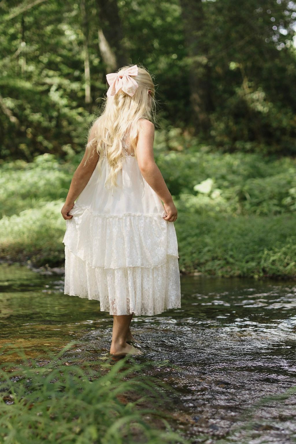 A young blonde girl in a white lace dress and pink bow wades barefoot in a peaceful forest stream.