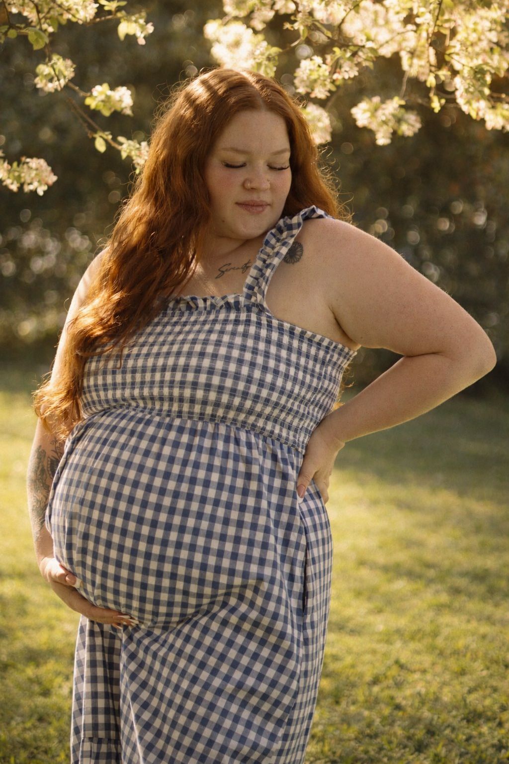 A pregnant woman in a blue gingham dress poses in golden sunlight with blooming cherry blossoms in the background.