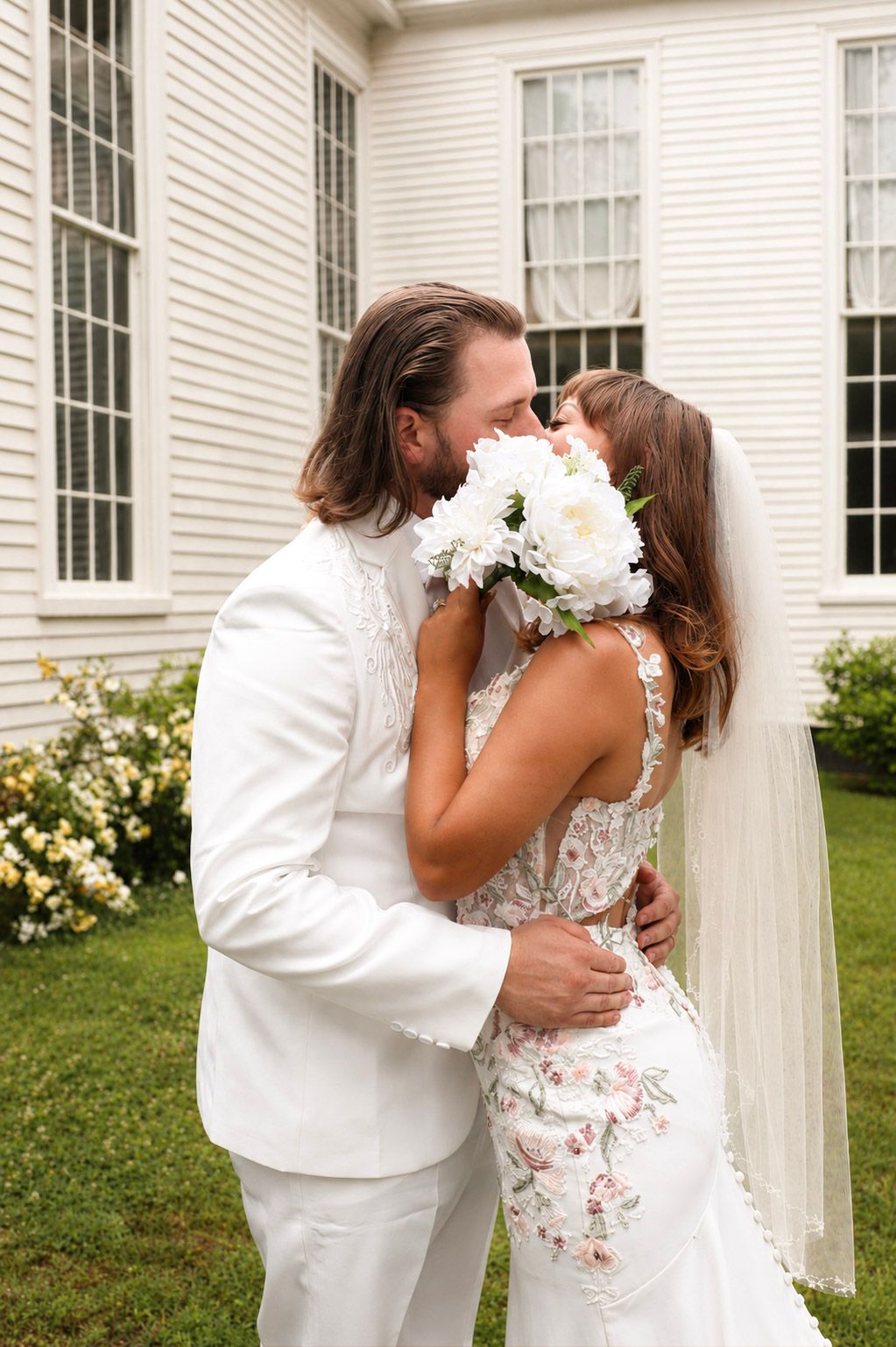 Bride and groom sharing a kiss behind white floral bouquet outside a white chapel on their wedding day.