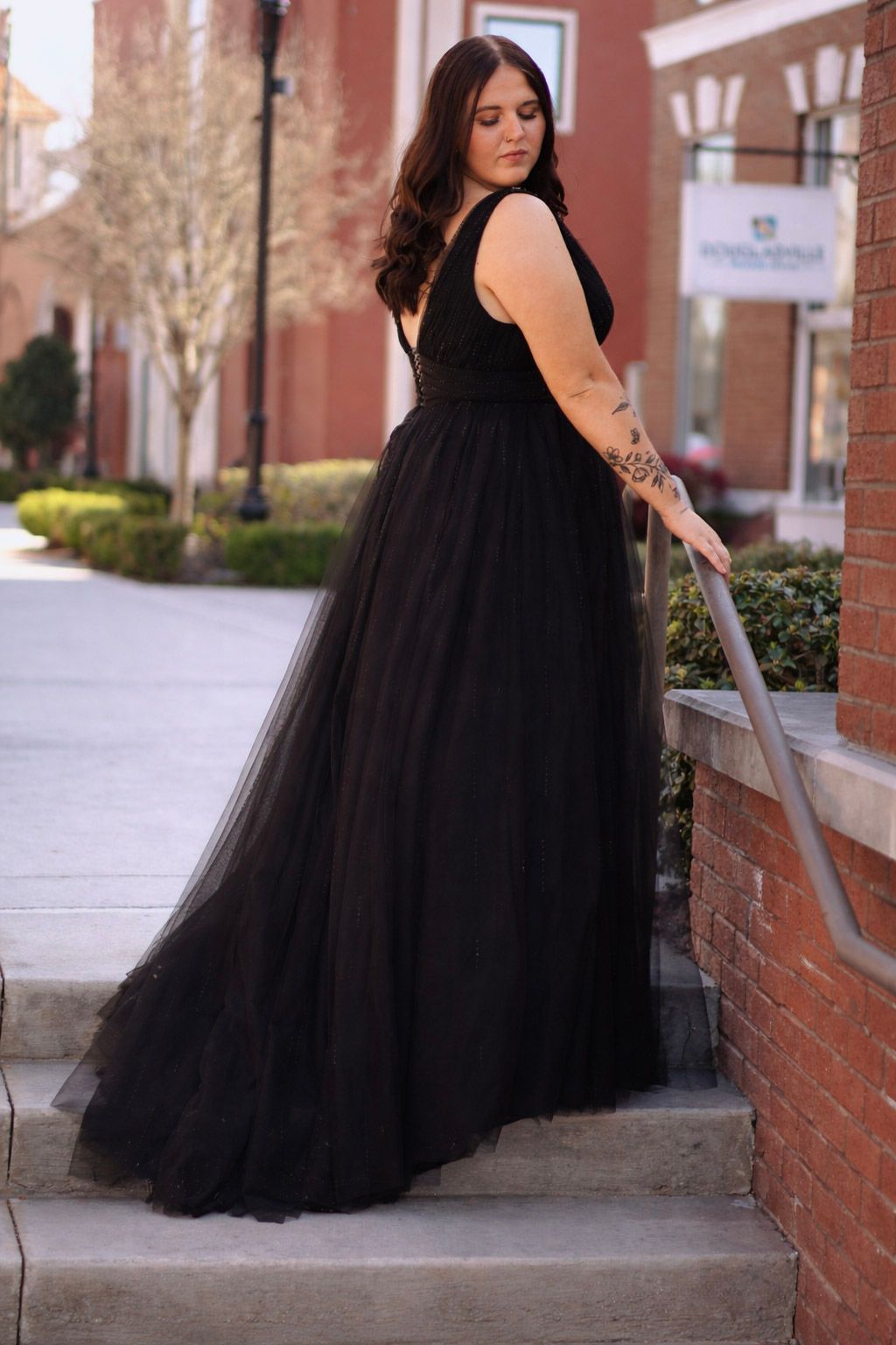 Woman in elegant black tulle ballgown poses on brick steps, showcasing flowing skirt and tattoo.