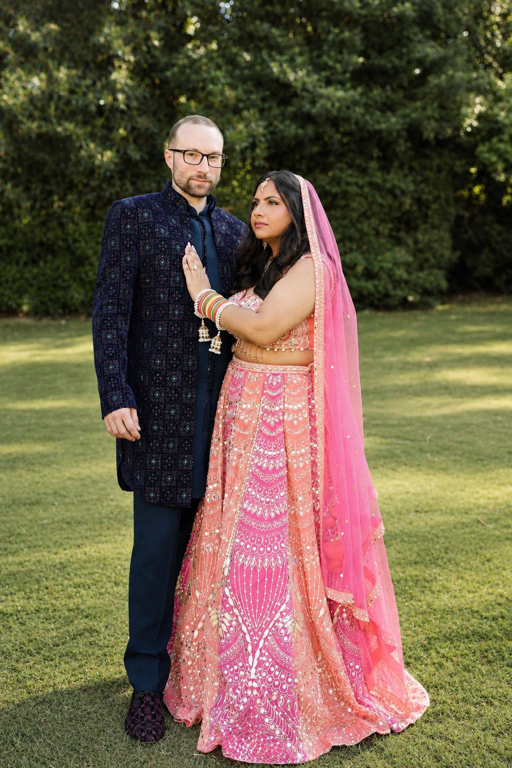 Couple posing outdoors in Indian wedding attire, woman in pink lehenga with dupatta, man in navy sherwani, lush green background.