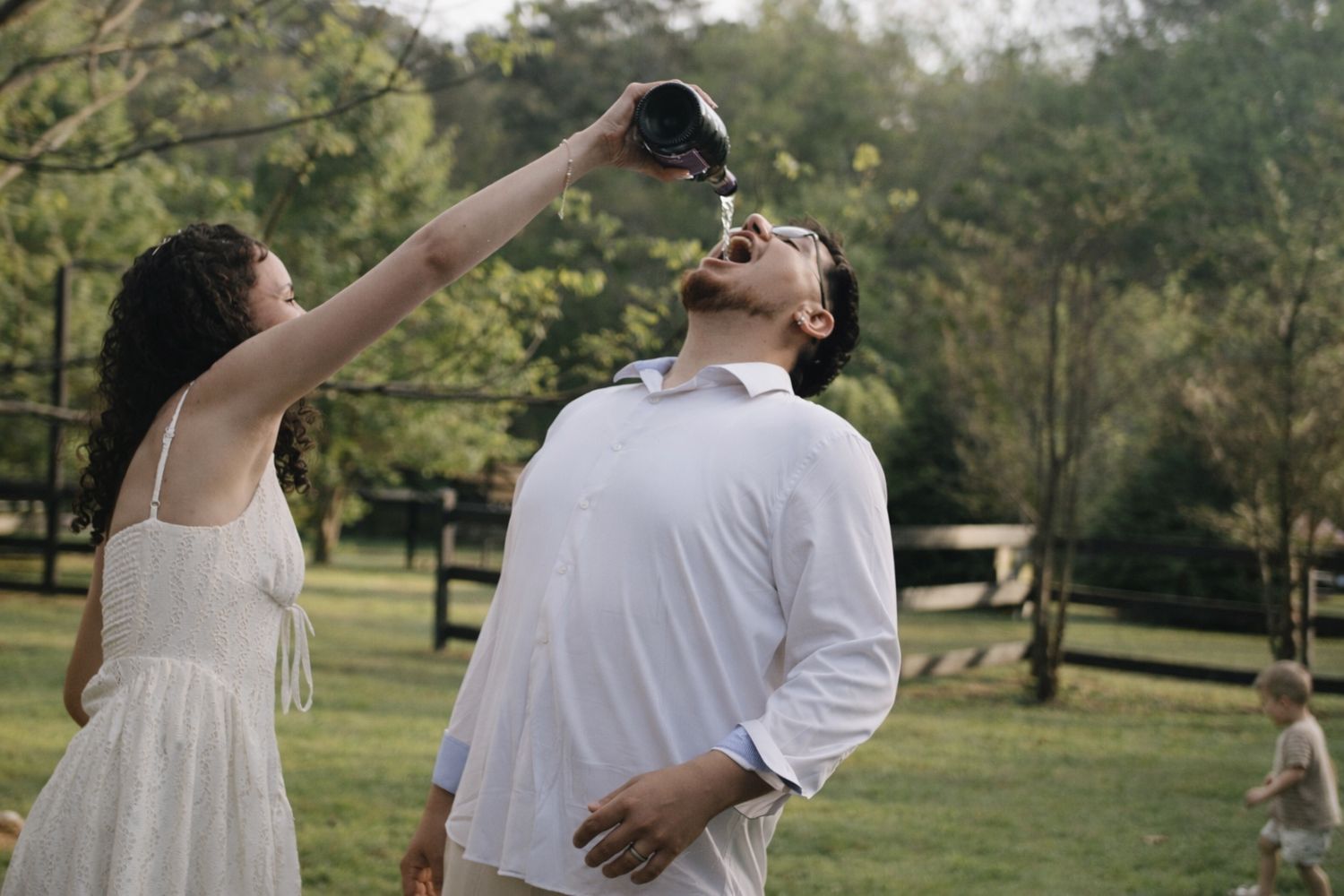 Woman in white dress pouring drink into mans mouth outdoors in a lush green park setting.