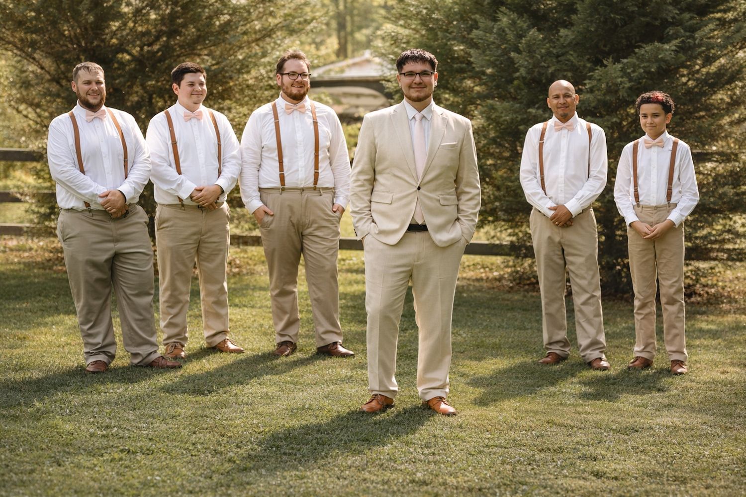 Groom stands in front of six groomsmen wearing tan suits with rust orange suspenders and bow ties on a green lawn.