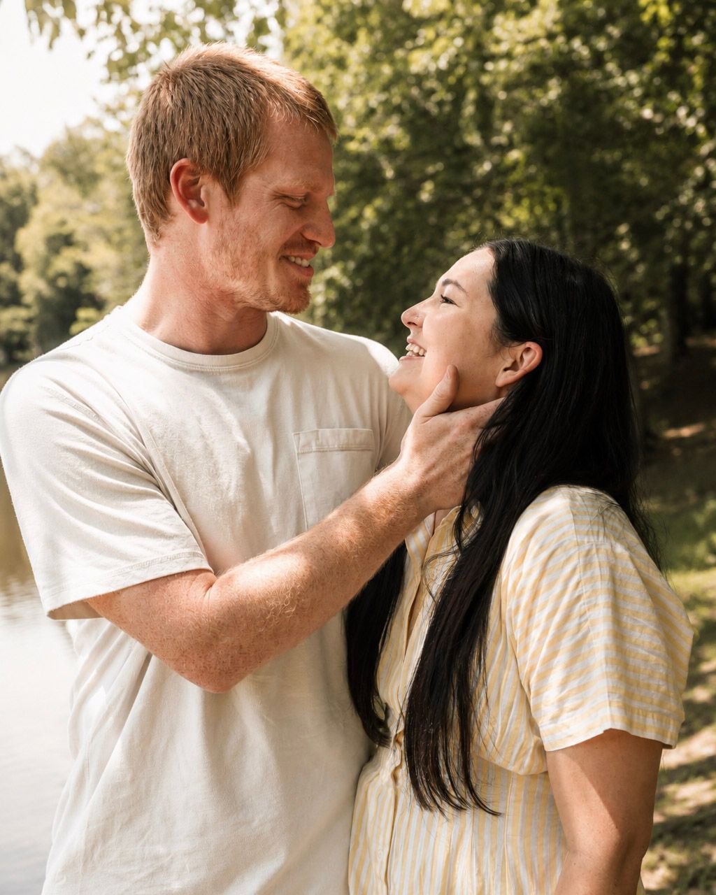 A smiling couple shares a tender moment outdoors, man gently holding womans face near a sunlit lake.