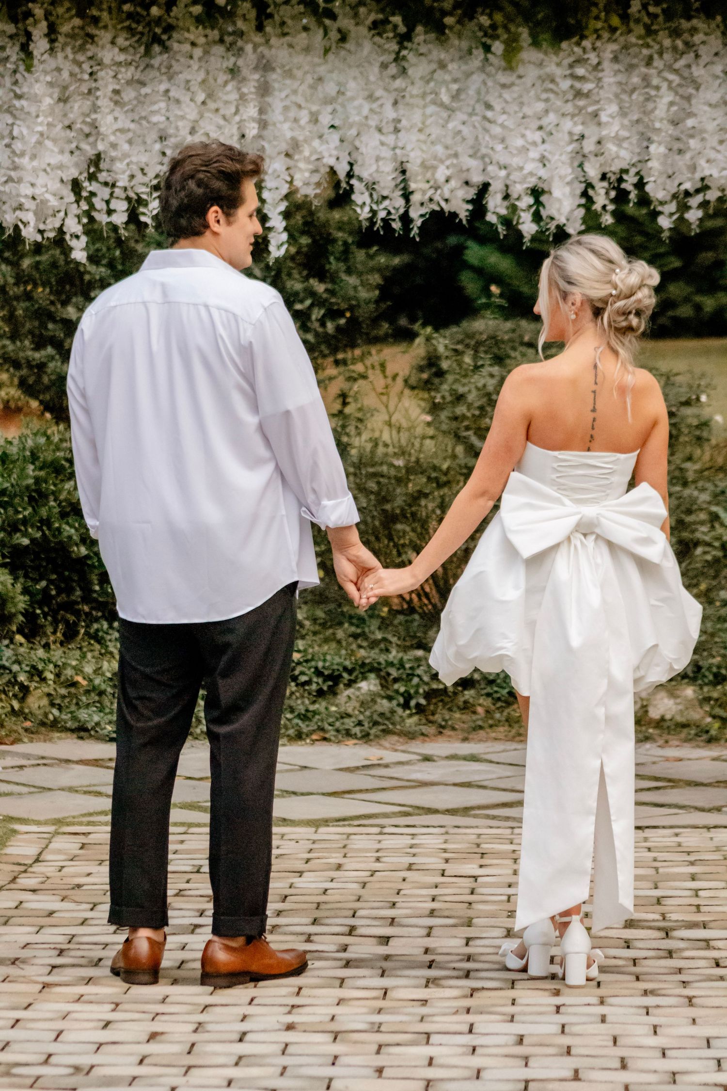 A couple in wedding attire walks hand in hand on a brick pathway.