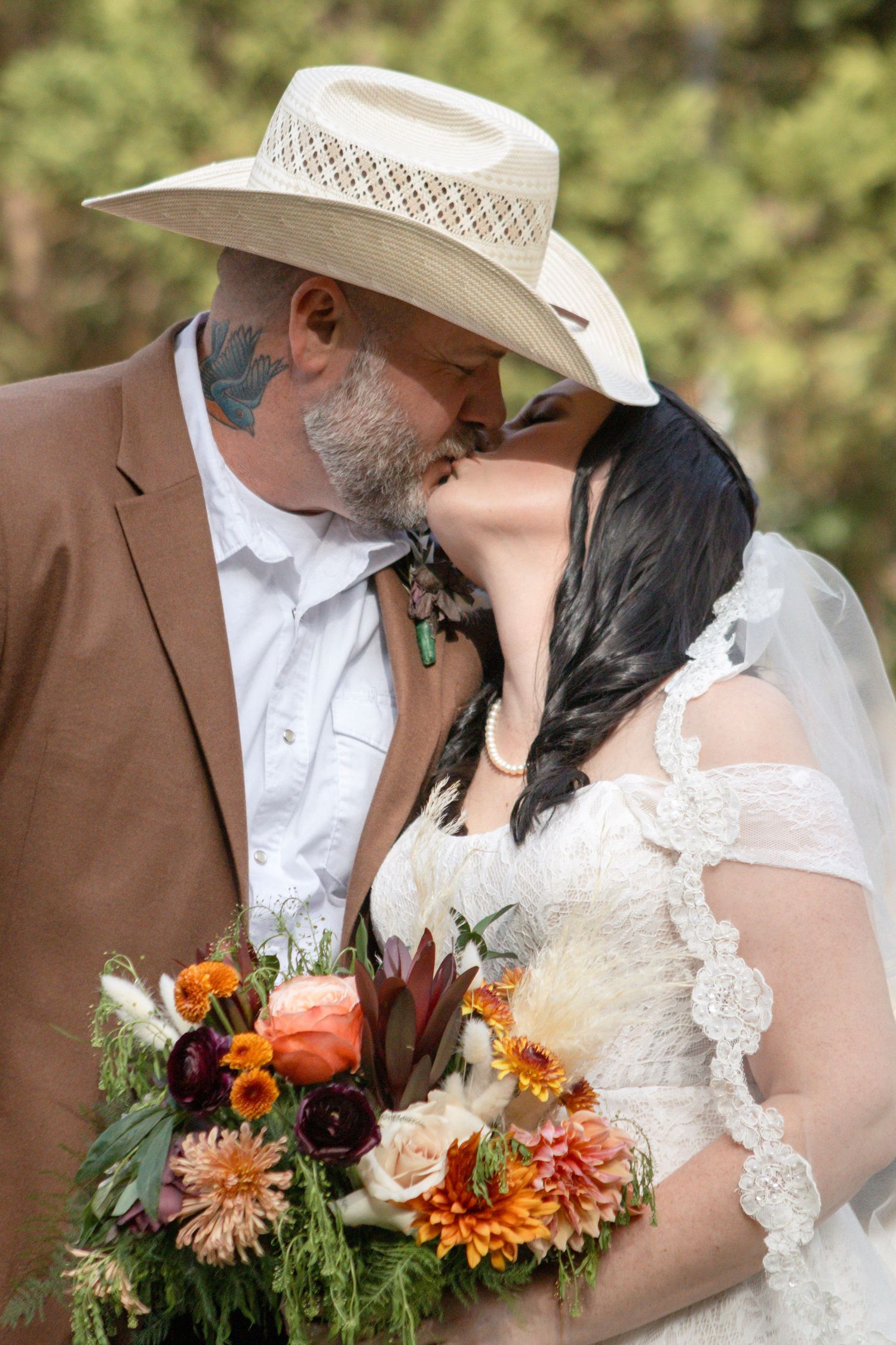 A romantic wedding couple shares a kiss while holding vibrant orange and burgundy fall bouquets in an outdoor setting.