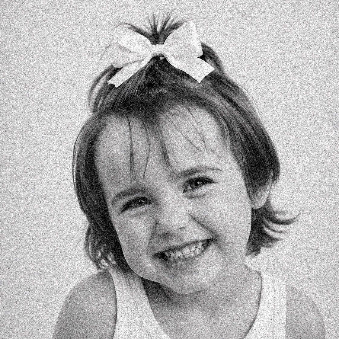Black and white portrait of a smiling toddler girl with a white bow in her hair wearing a tank top.