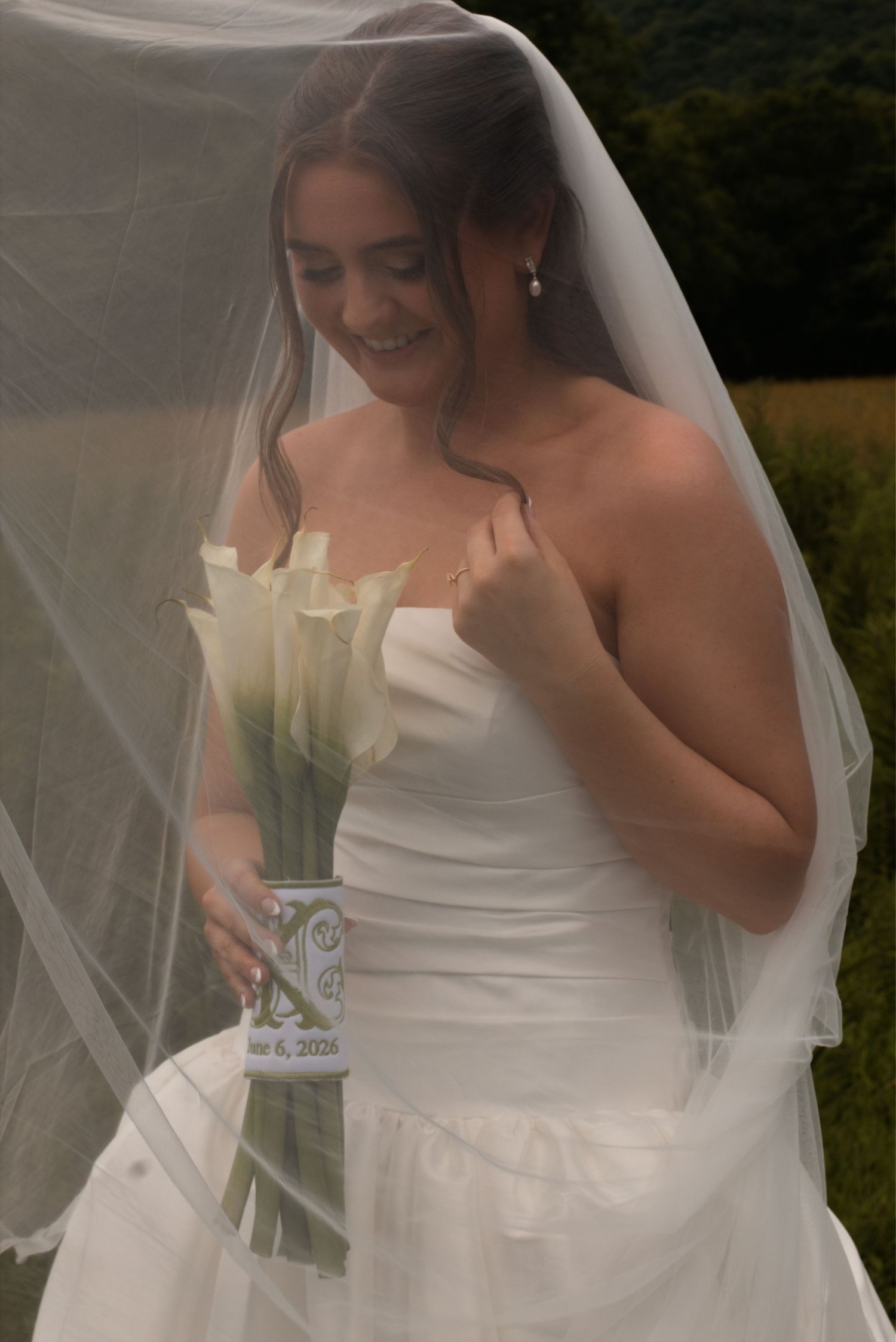 Smiling bride in strapless white gown and veil holds white calla lily bouquet outdoors on her wedding day.