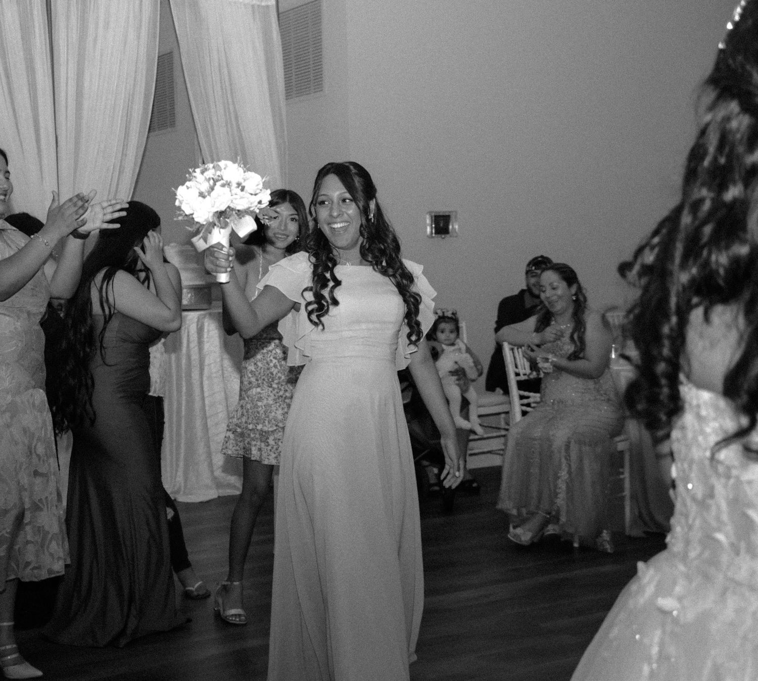 Bride in white gown tossing bouquet to excited bridesmaids at indoor wedding reception in black and white photo.