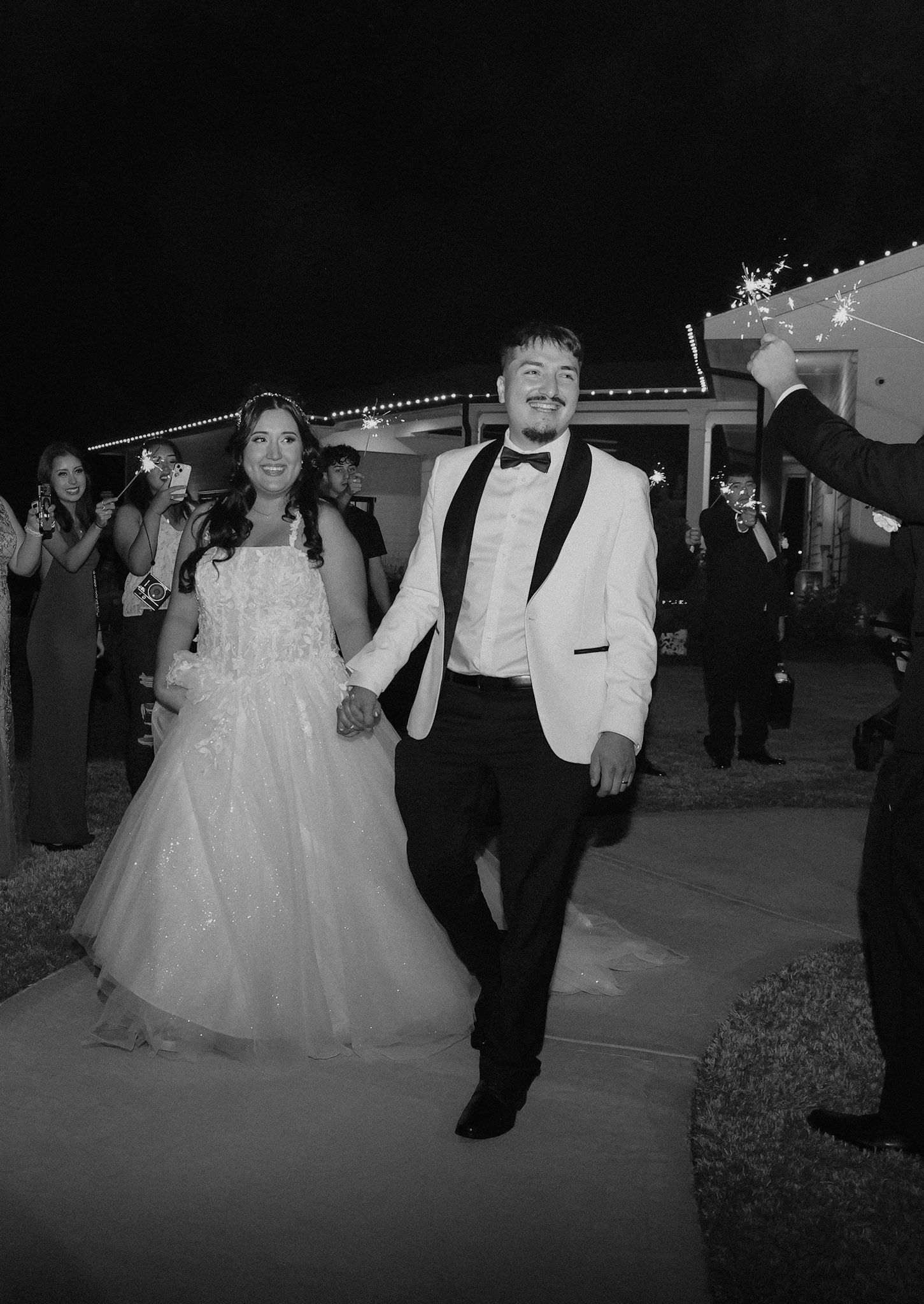 Black and white photo of a bride and groom holding hands, walking joyfully at their outdoor wedding reception.