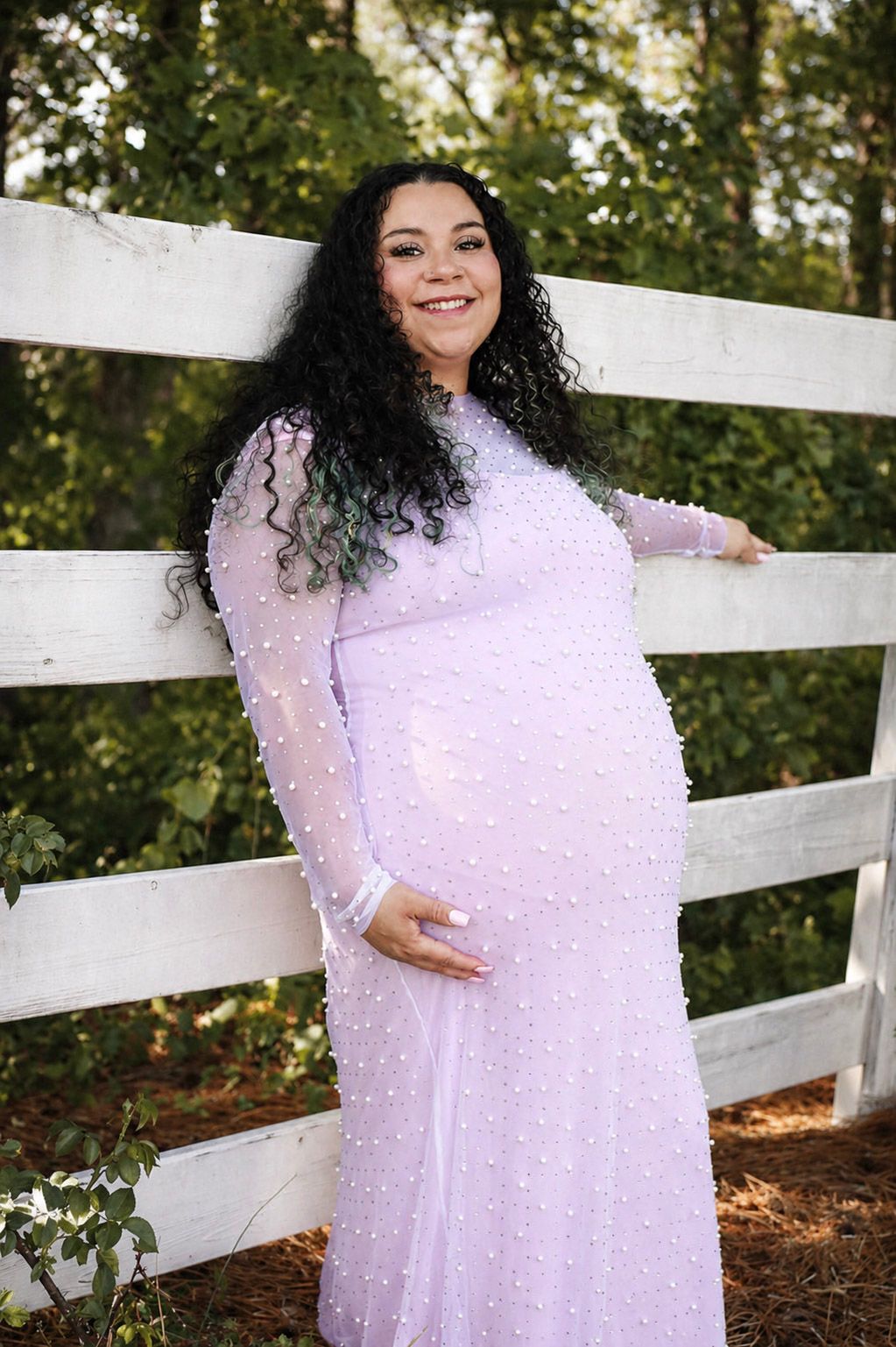 Pregnant woman in lavender pearl gown smiling by white fence outdoors.