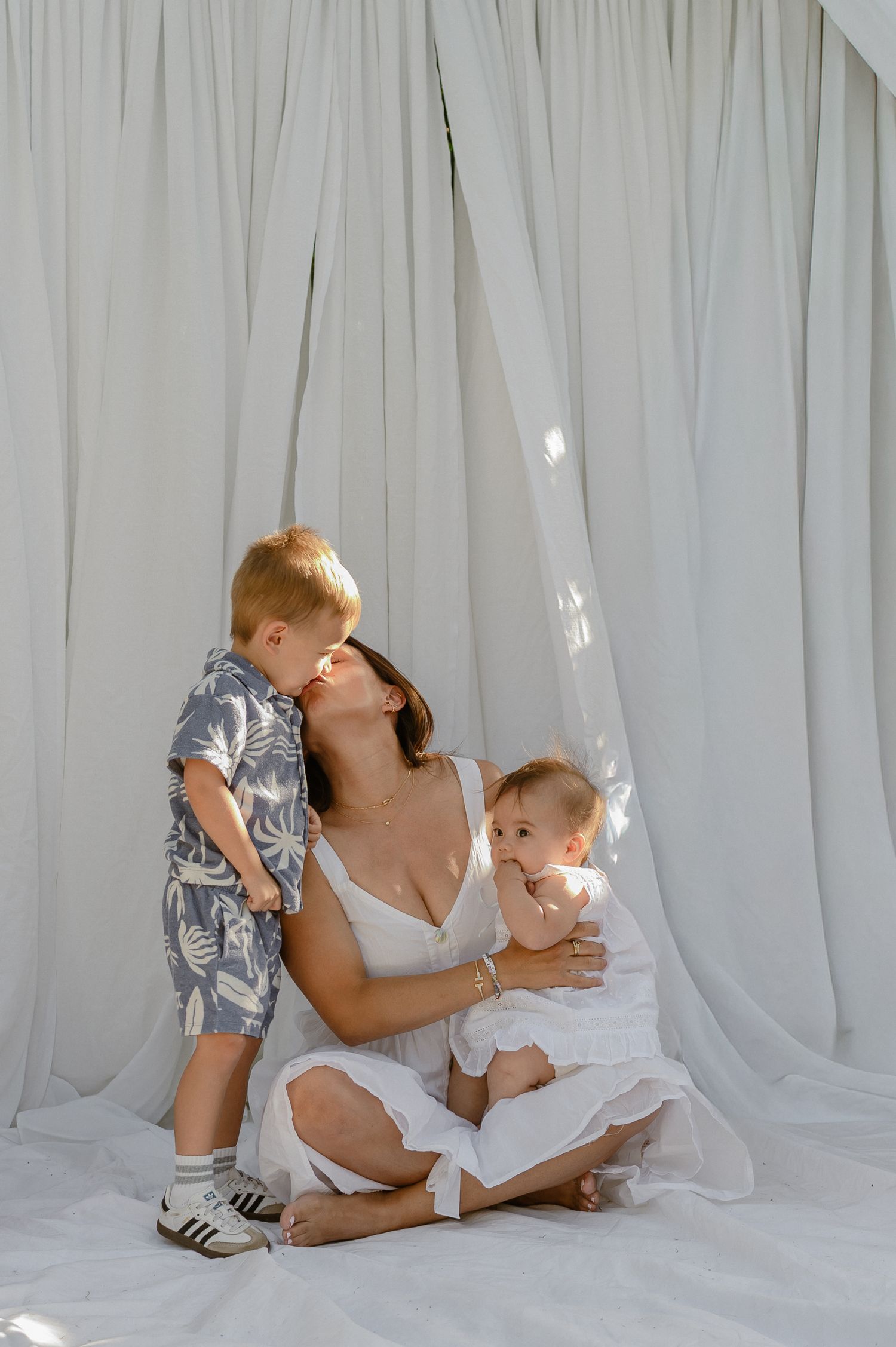 Mother in white dress sitting with toddler son and baby in front of flowing white curtains in natural light.