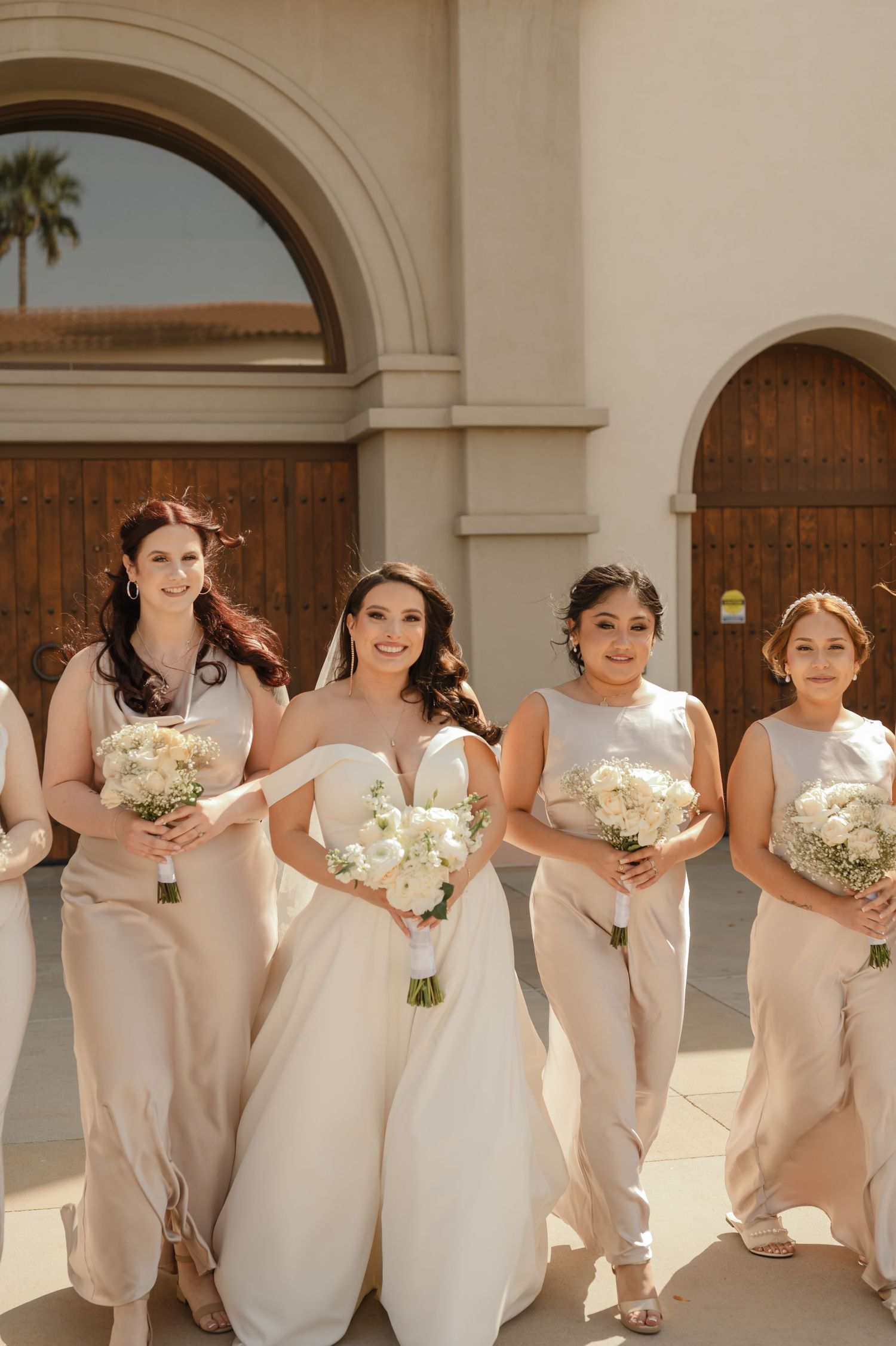 Bride and bridesmaids in champagne gowns holding white bouquets, walking outside mission-style venue.
