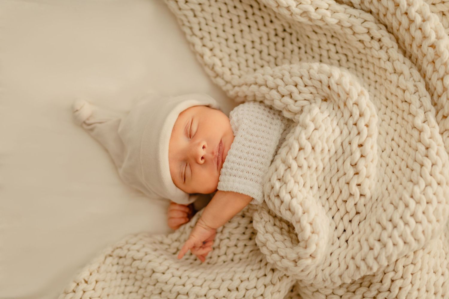A newborn sleeps peacefully in a chunky cream knit blanket, captured during an in home lifestyle newborn session.