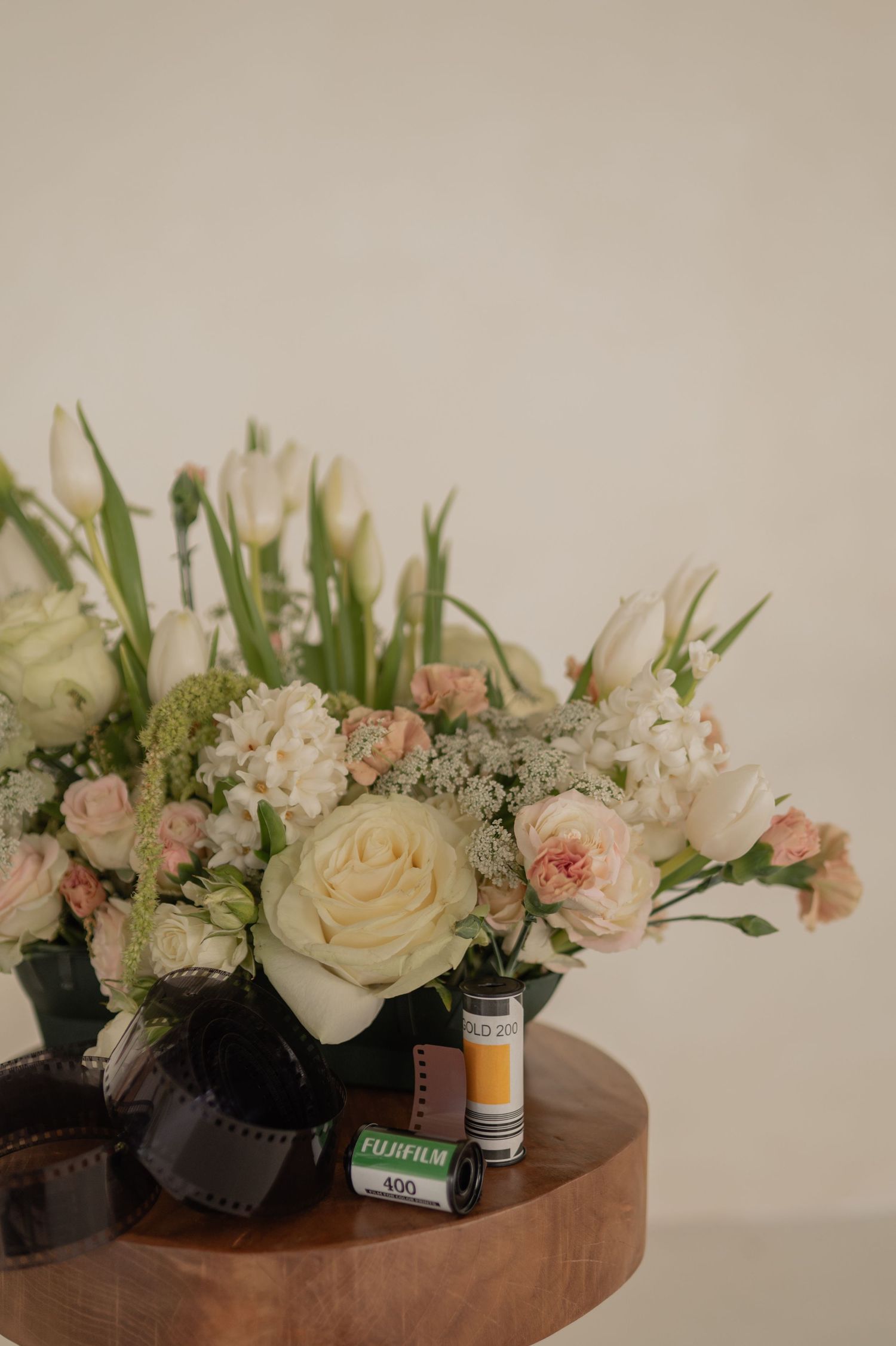 A floral arrangement of white roses, tulips, and pink blooms sits beside a roll of Fujifilm 400 on a wooden table.