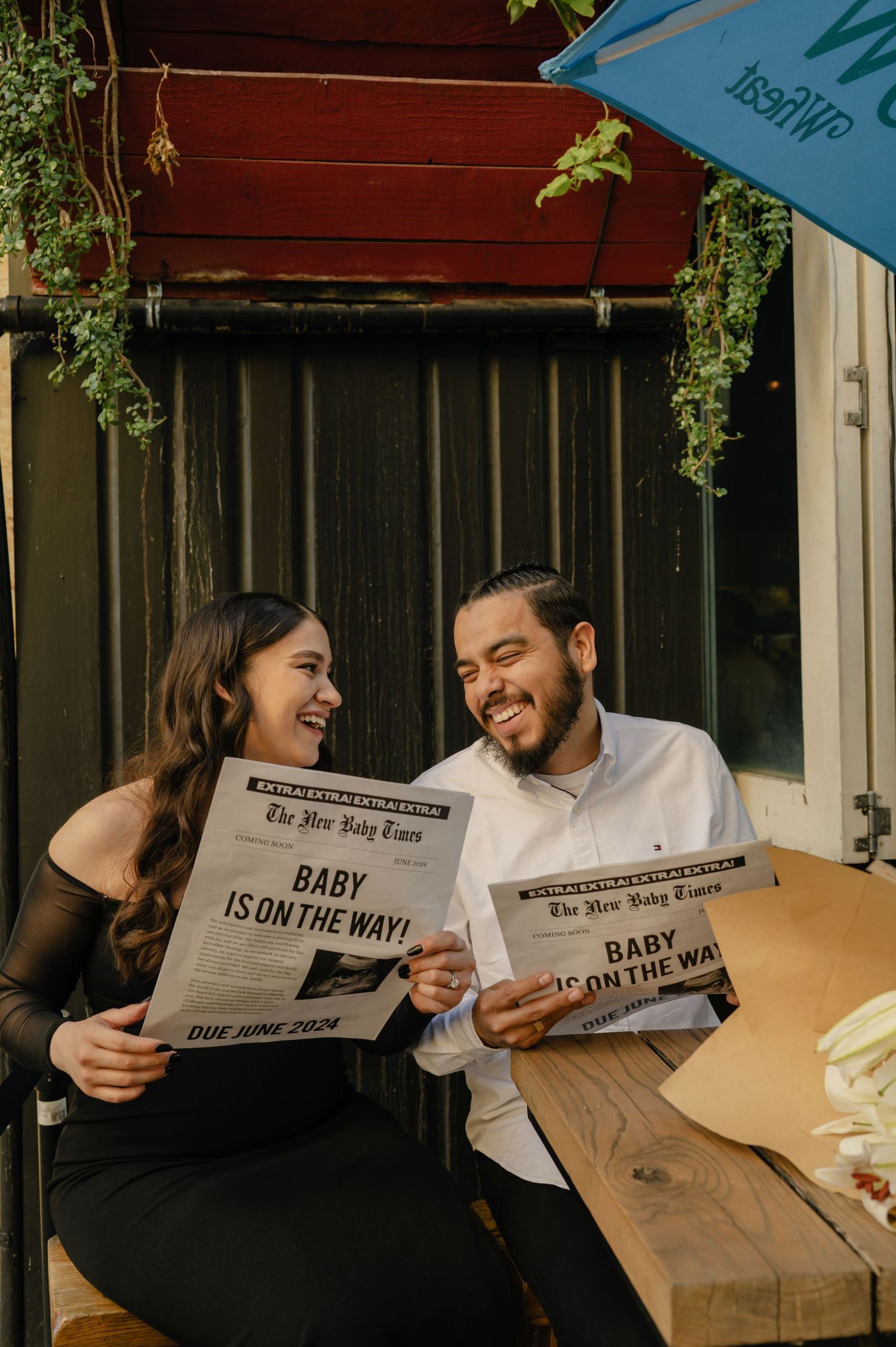 A laughing couple sits at an outdoor table holding newspapers with Baby Is On The Way headlines announcing their June 2024 pregnancy.