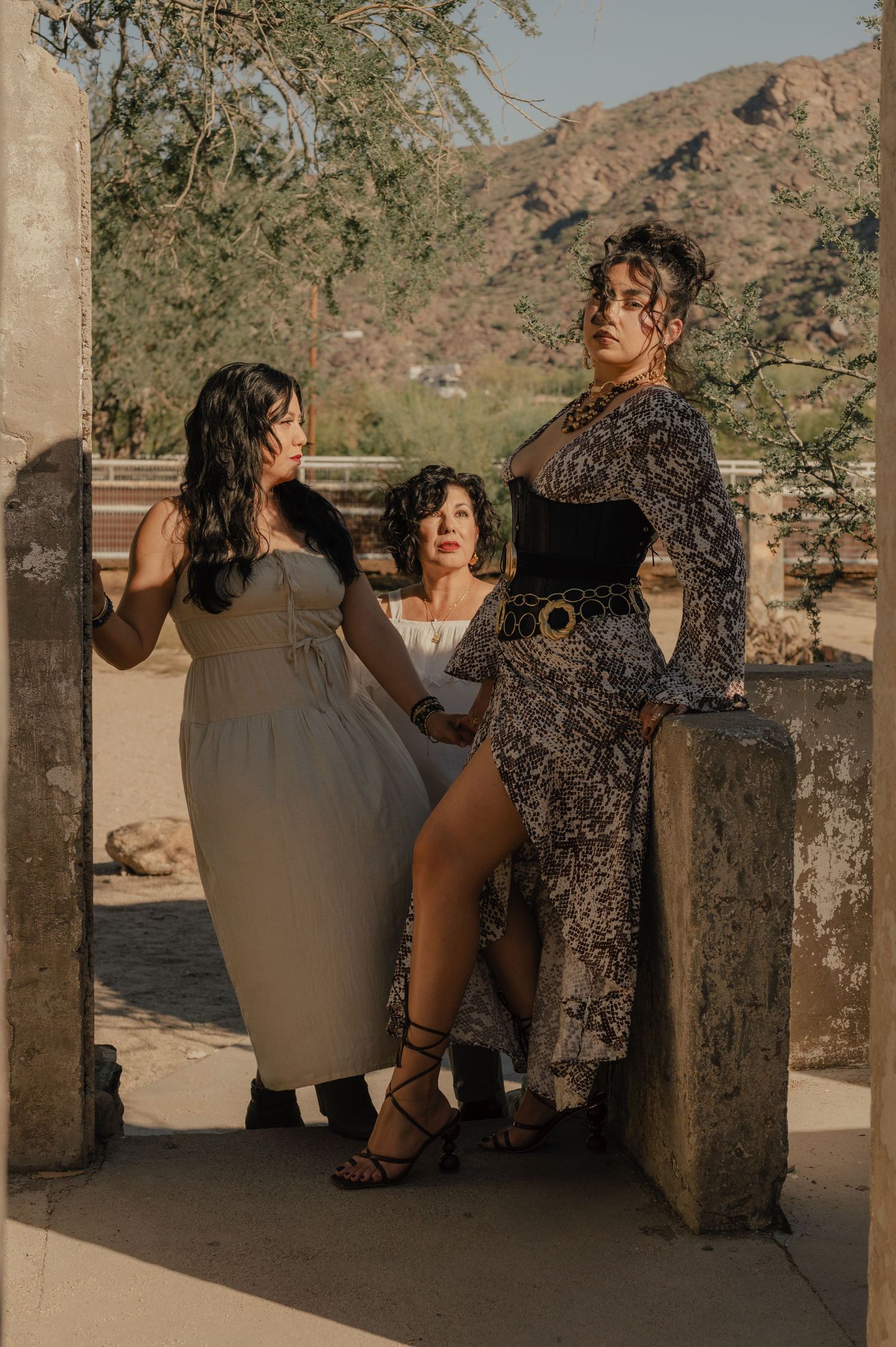 Group of three women pose together in the golden hour light by a stone wall and desert backdrop.