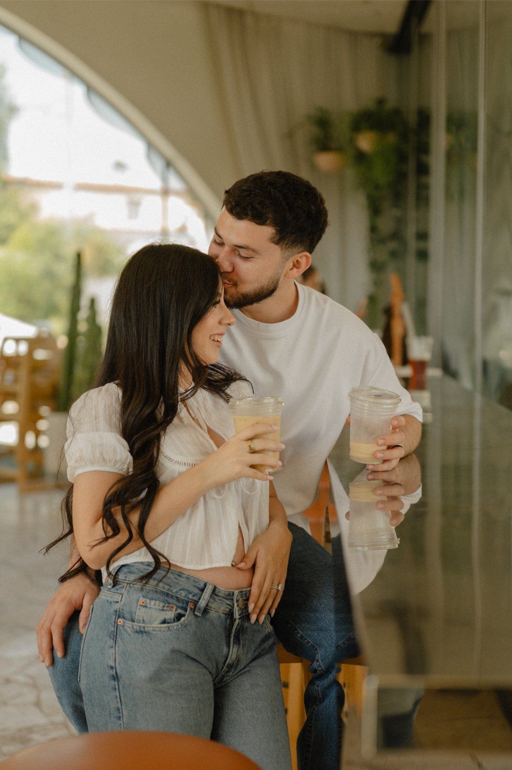 Couple embracing with a forehead kiss while holding their iced coffees at Sophia's Brunch Counter for their pregnancy announcement session