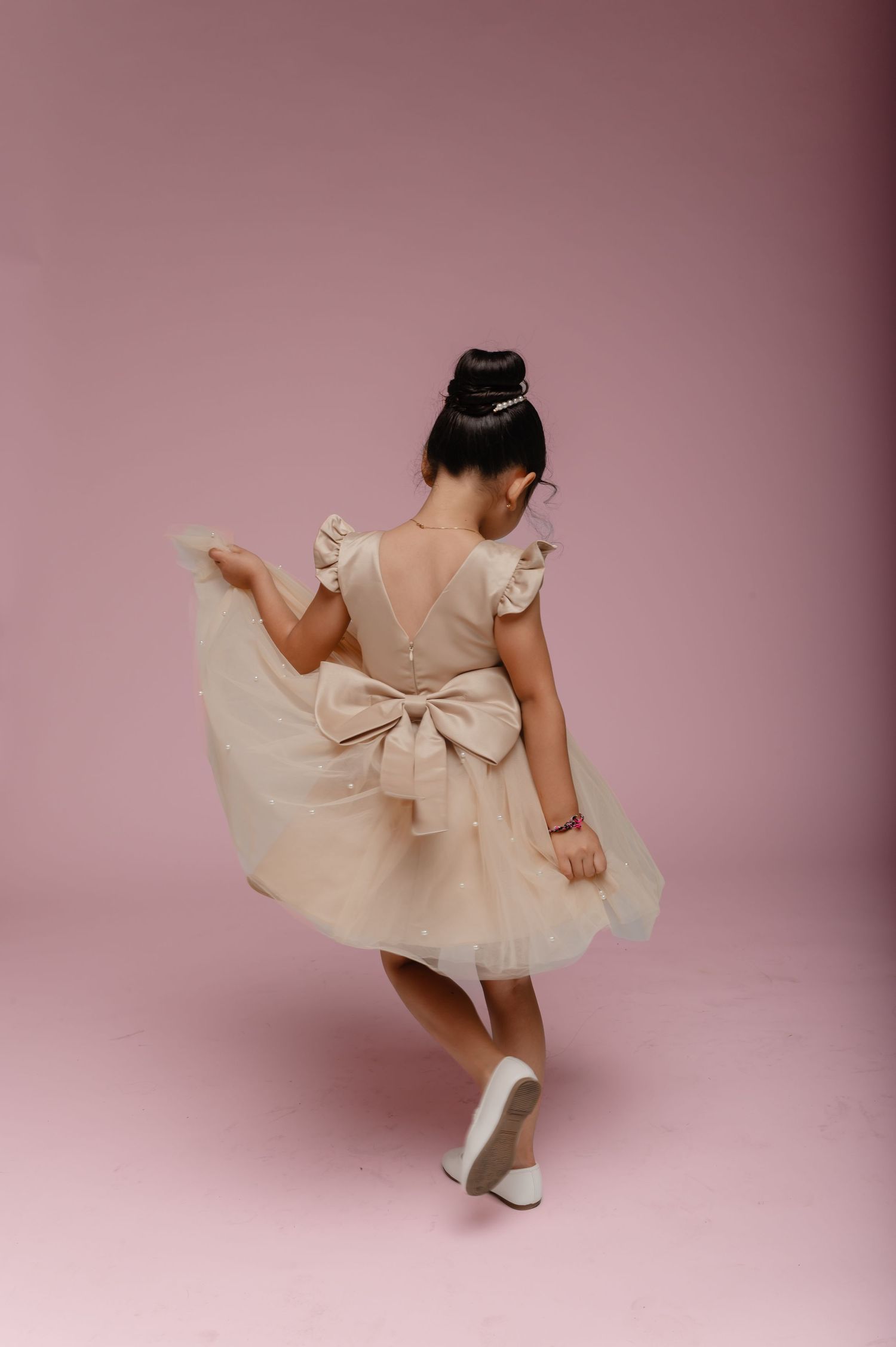 Young girl in a beige tulle dress with a bow twirling gracefully against a soft pink studio background.