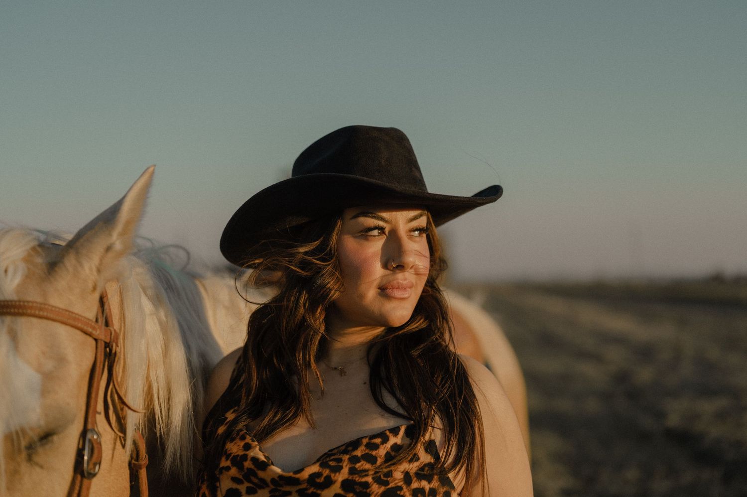 A woman in a leopard print top and black cowboy hat poses beside a white horse at sunset.