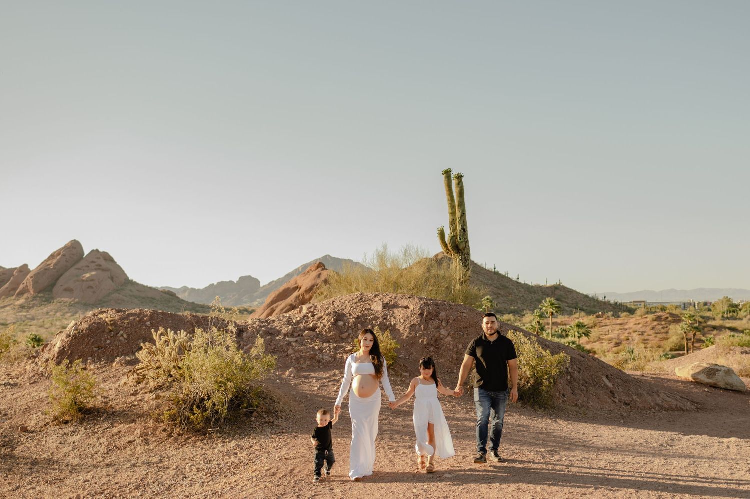 Family of four walks hand-in-hand through Arizona desert landscape with saguaro cactus and red rock formations.