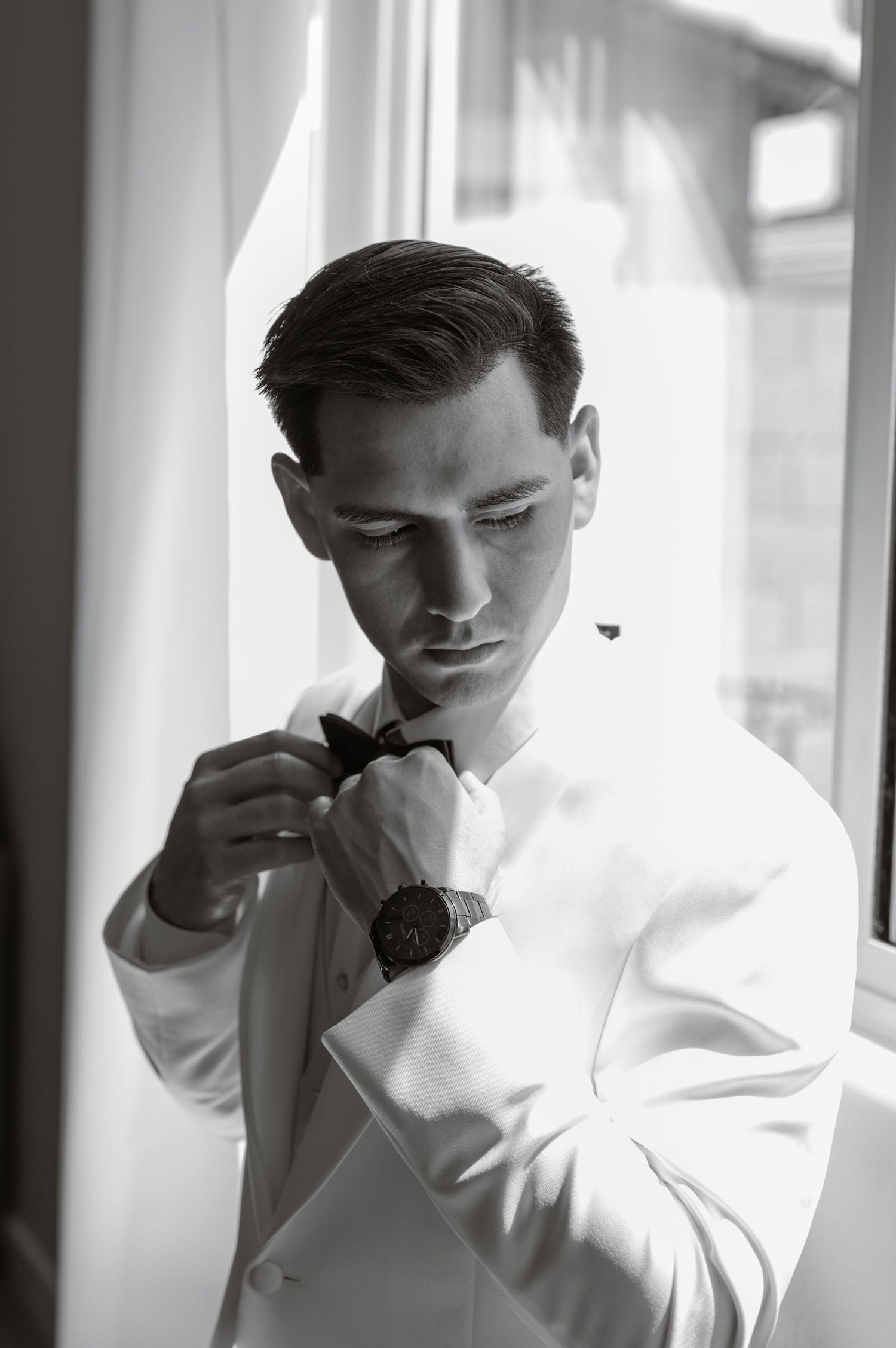 Groom adjusts cufflinks while standing by window in black-and-white portrait.