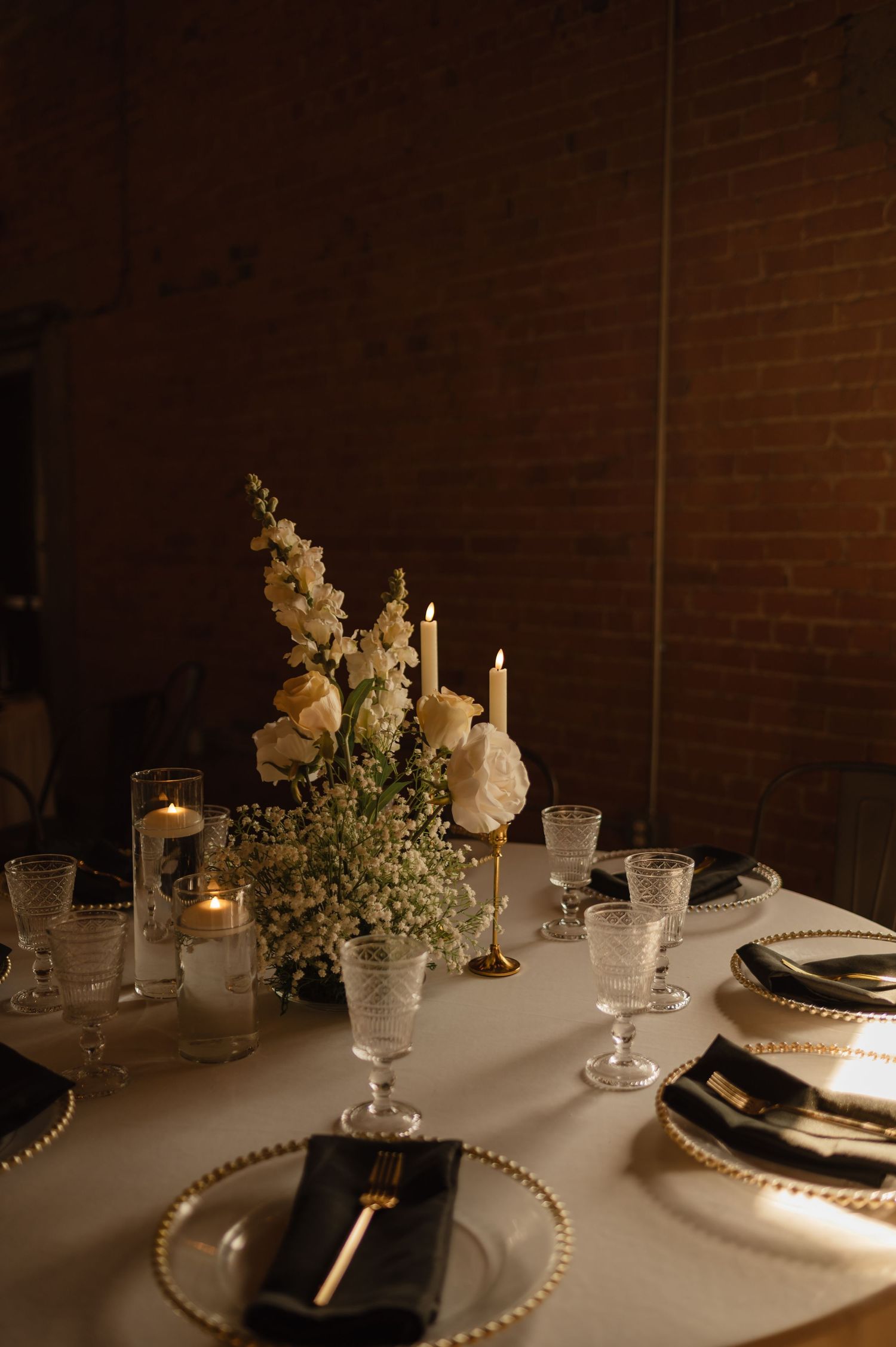 Elegant wedding reception table with black and gold place settings, candles, and white floral centerpiece.