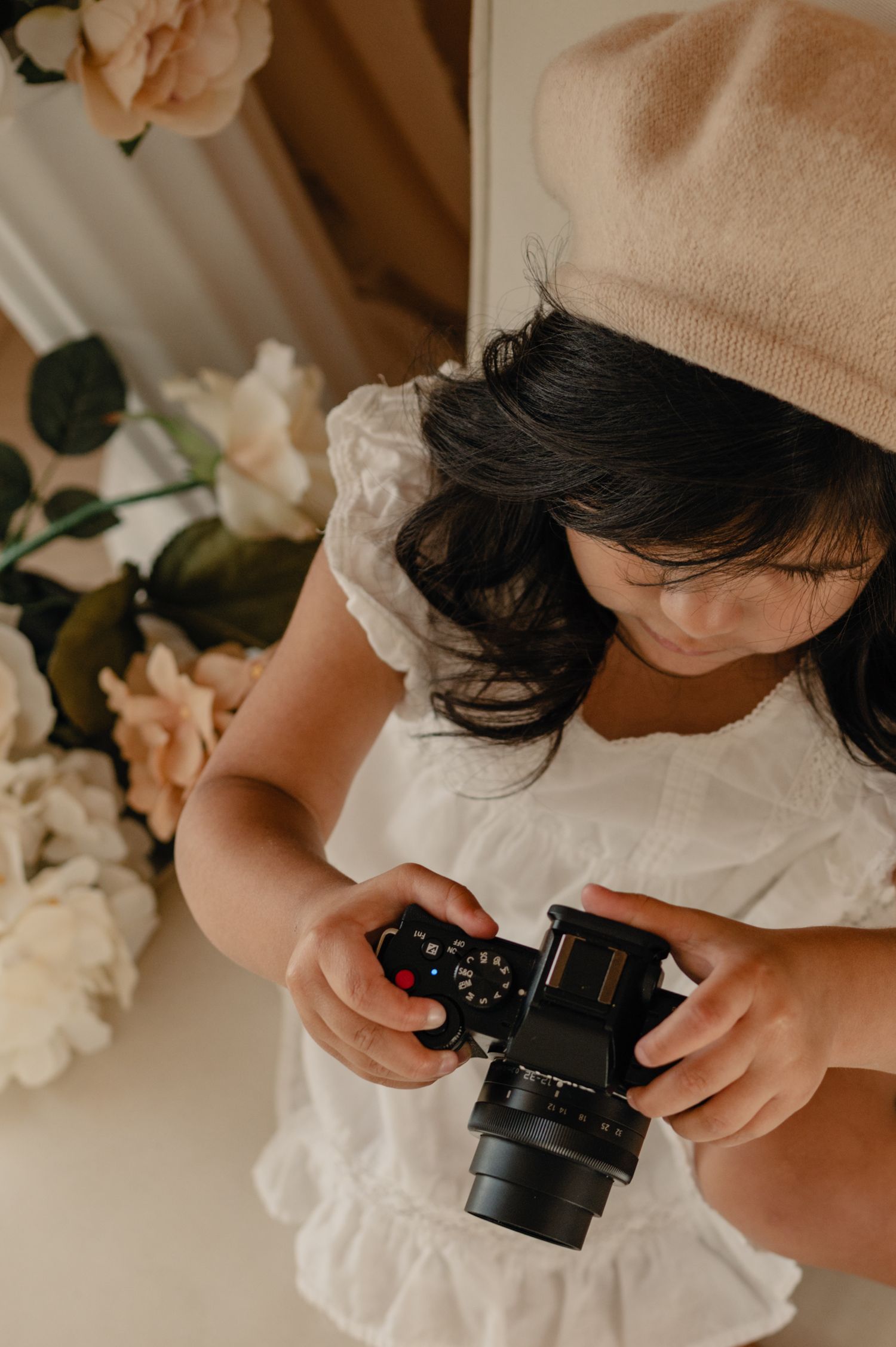 Candid moment of a young girl holding a camera, styled in a neutral setting with florals and natural light.