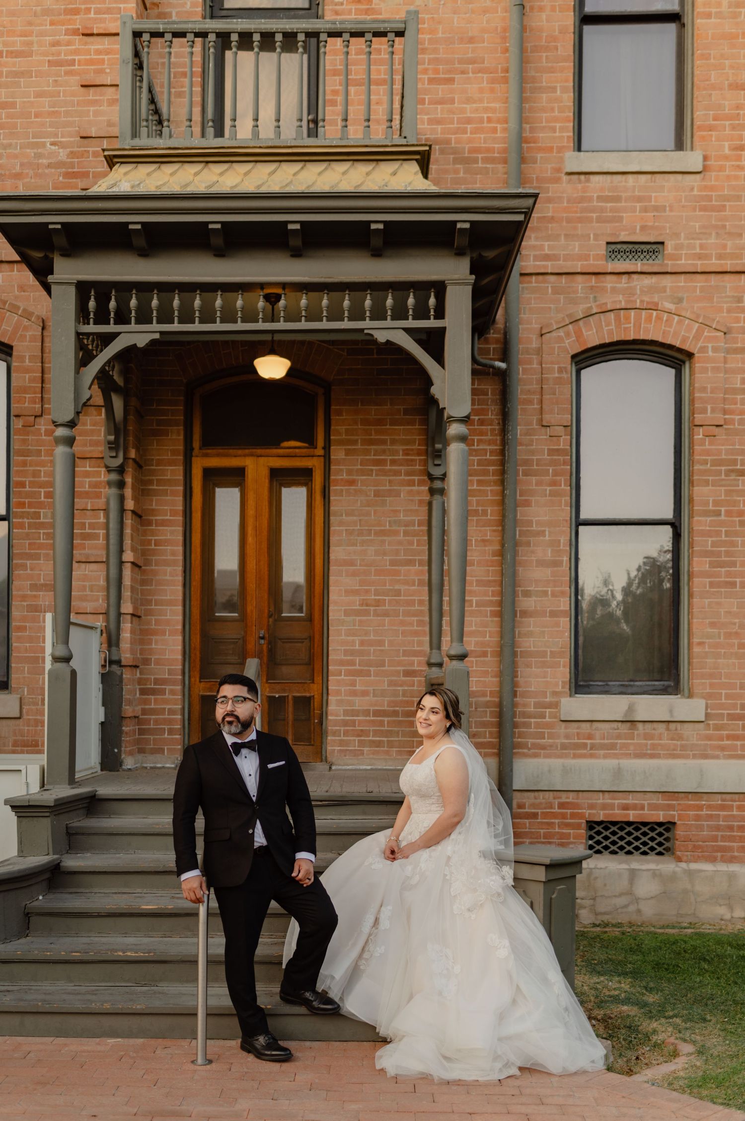 Bride and groom pose on steps of historic brick building, she in layered white gown, he in black tuxedo with bow tie.