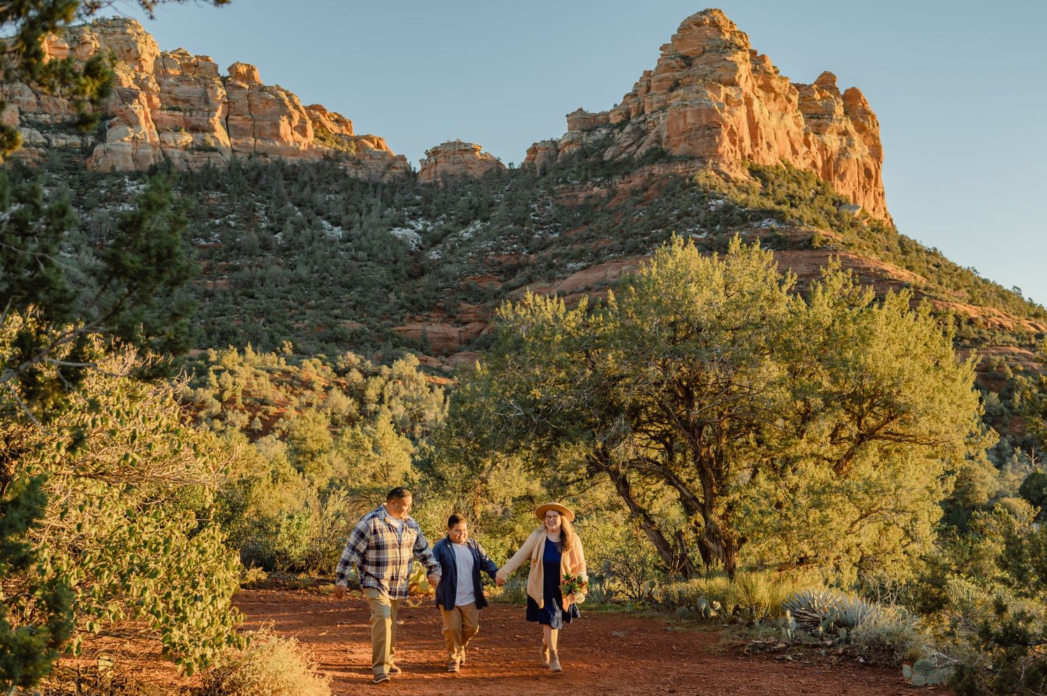 A family of three walks hand in hand along a red dirt trail beneath Sedona's iconic red rock formations.