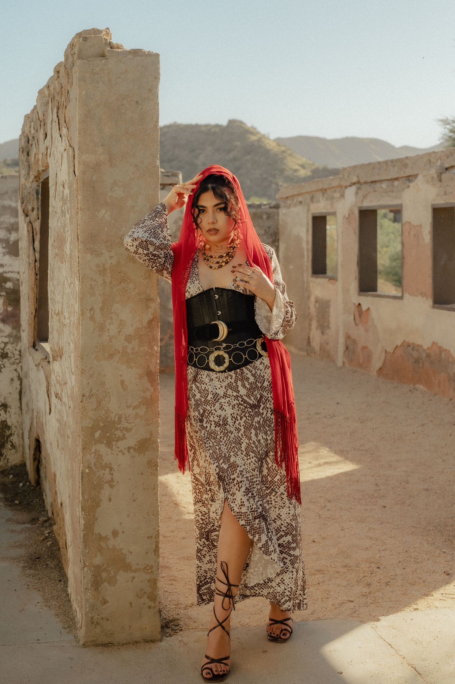 Woman in a red scarf and animal print dress stands in the ruins of a stone building with mountains in the background.